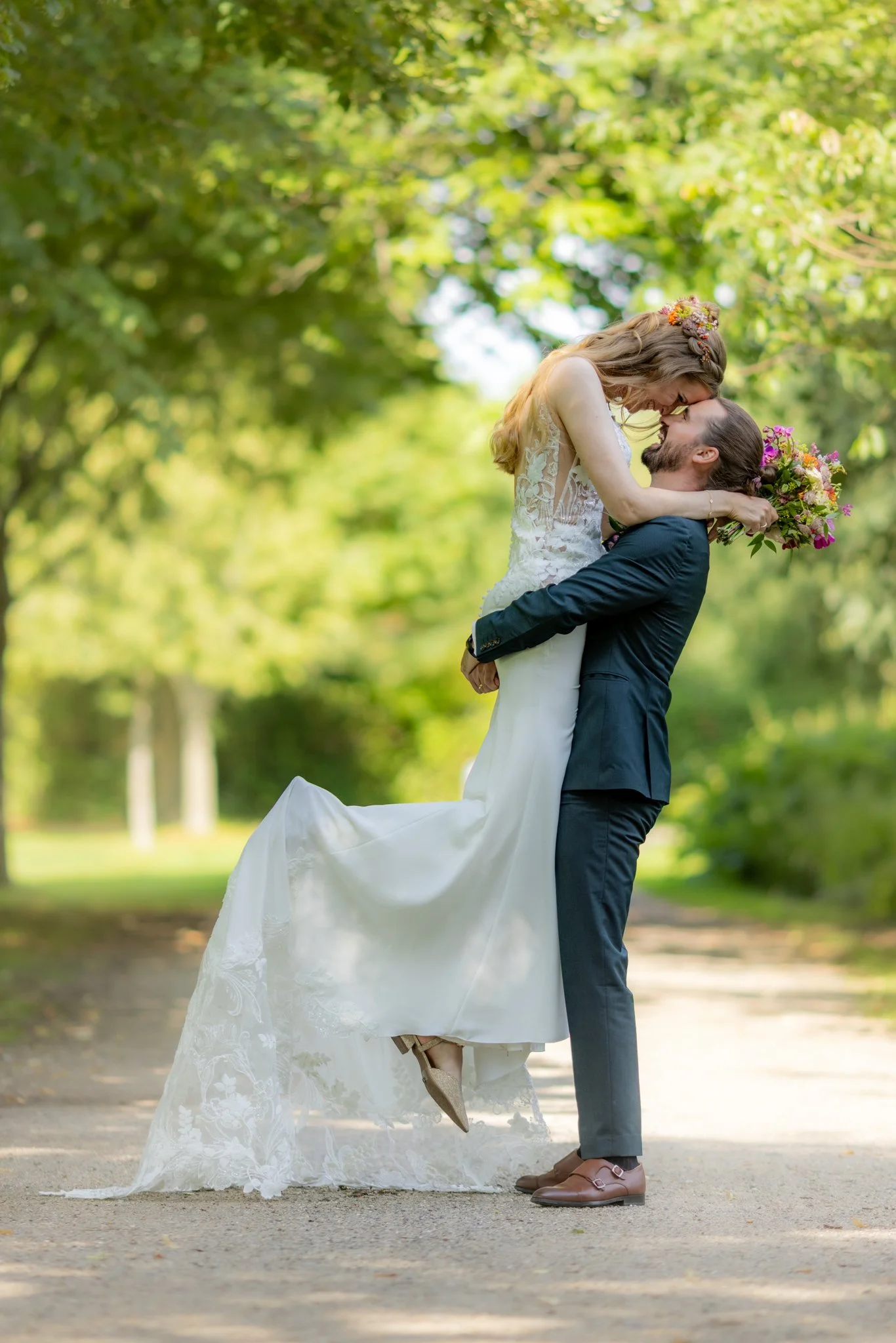 Et brudepar i en park, hvor bruden sidder på mandens arme, og de smiler til hinanden, omgivet af grønne træer.