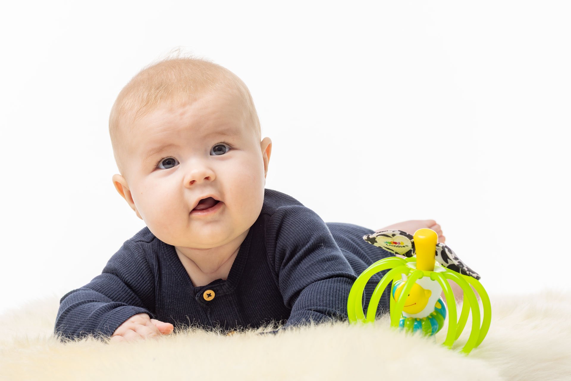 Klemt baby i navyblå body ligger på mødregræs med en legetøjs-rasle med grønne strimler og en butterfly på.