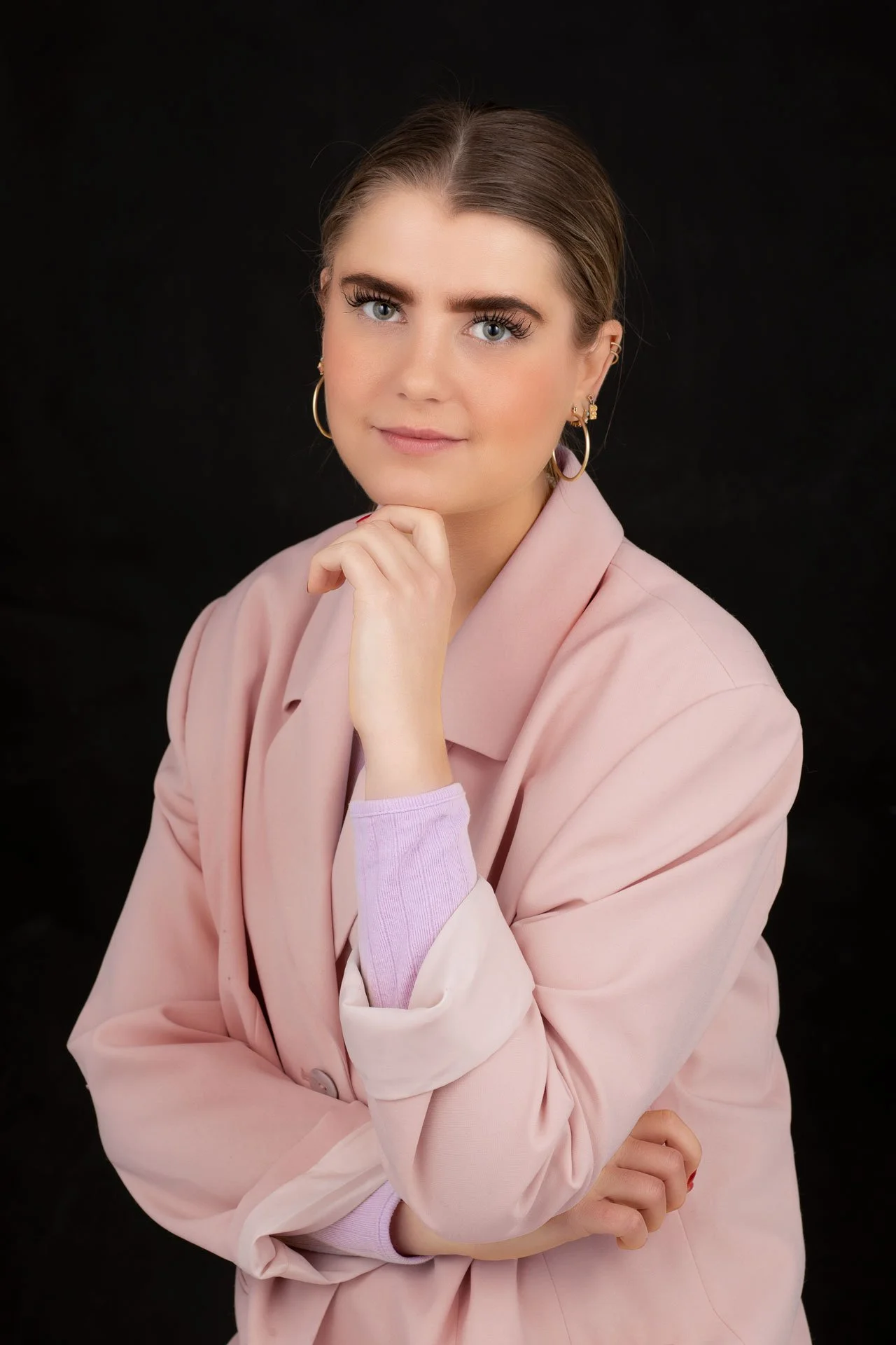 Portrait af en ung kvinde med lyst hår, blå øjne, iført en lyserosa blazer og blomstrede øreringe, mod en sort baggrund.