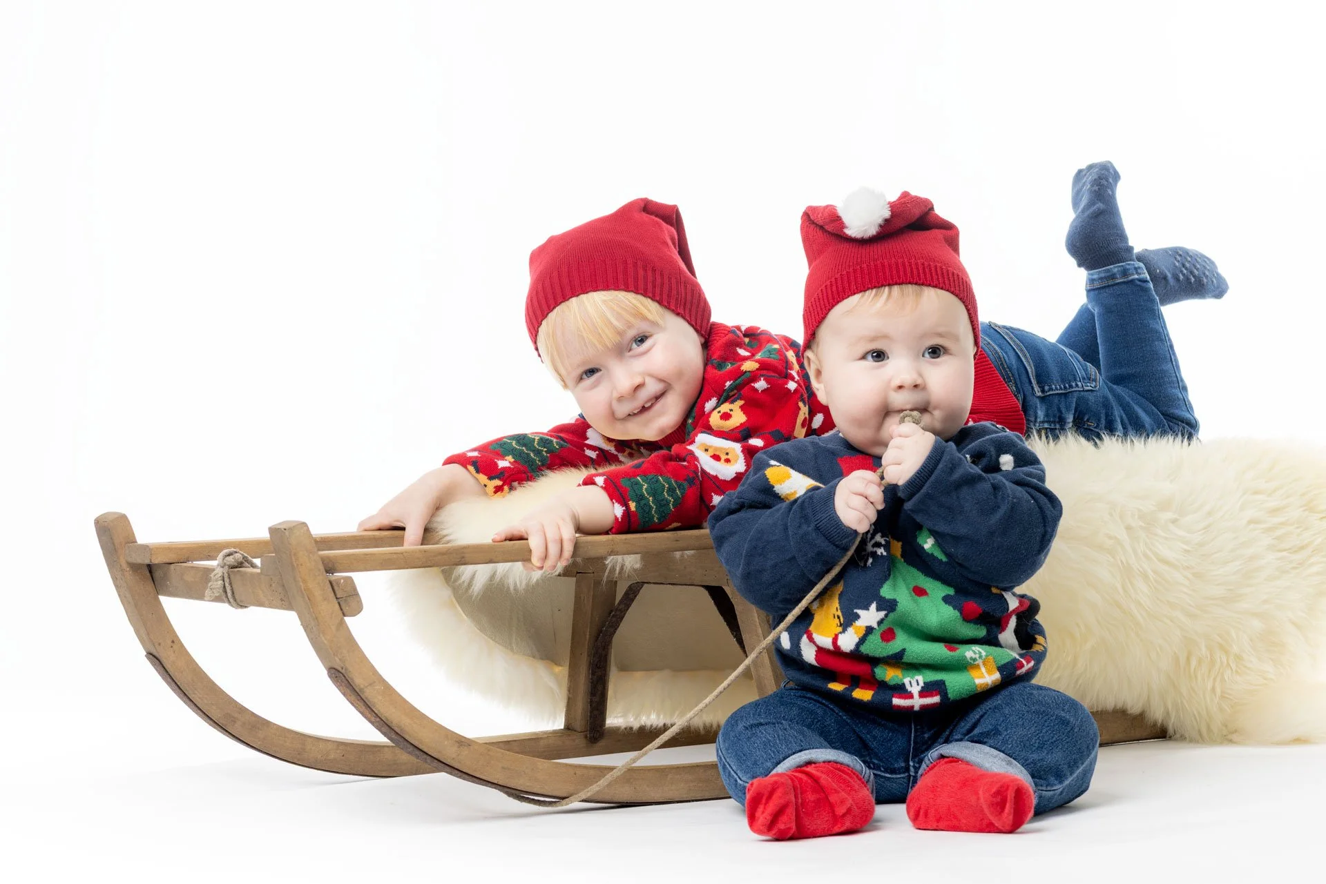 To børn i julegensæt med nisshuer, en sidder i skøjteslæde og en sidder på gulvet med en gave, begge smiler.