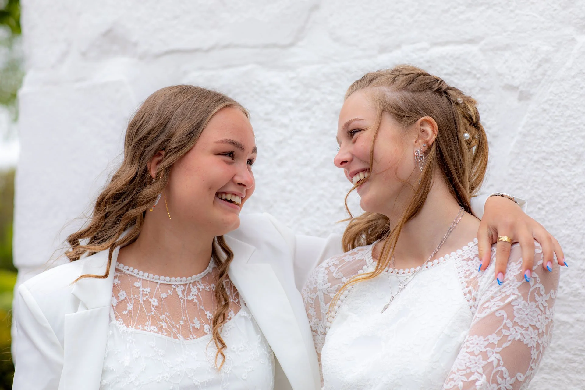 To unge kvinder smiler til hinanden og har armene omkring hinanden, begge i hvide kjoler, foran en hvid væg.