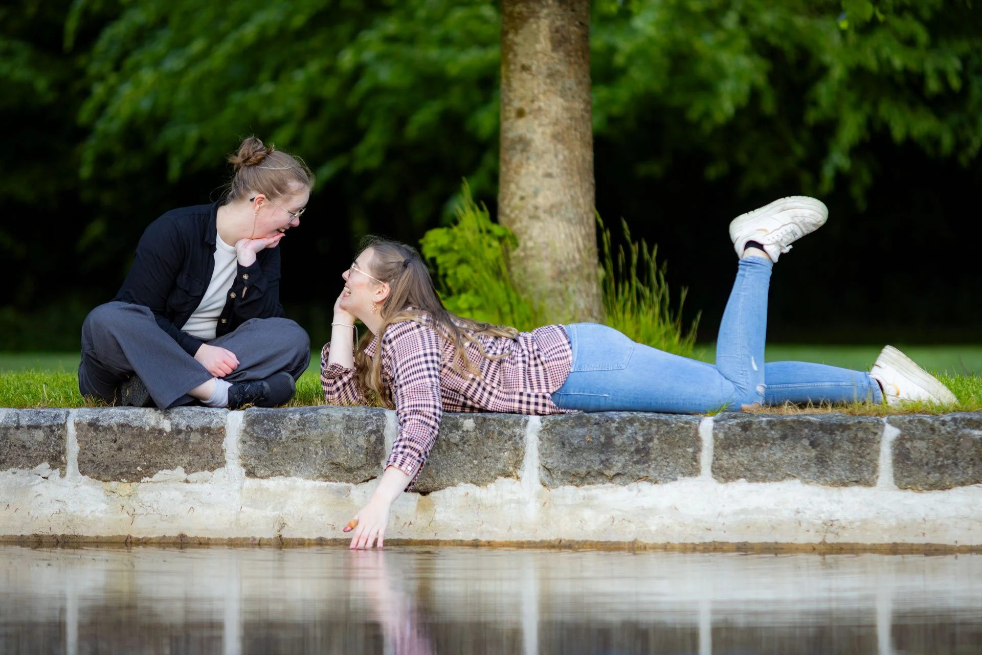 To unge kvinder, der leger ved en vandkant i en park, omgivet af grønne træer.