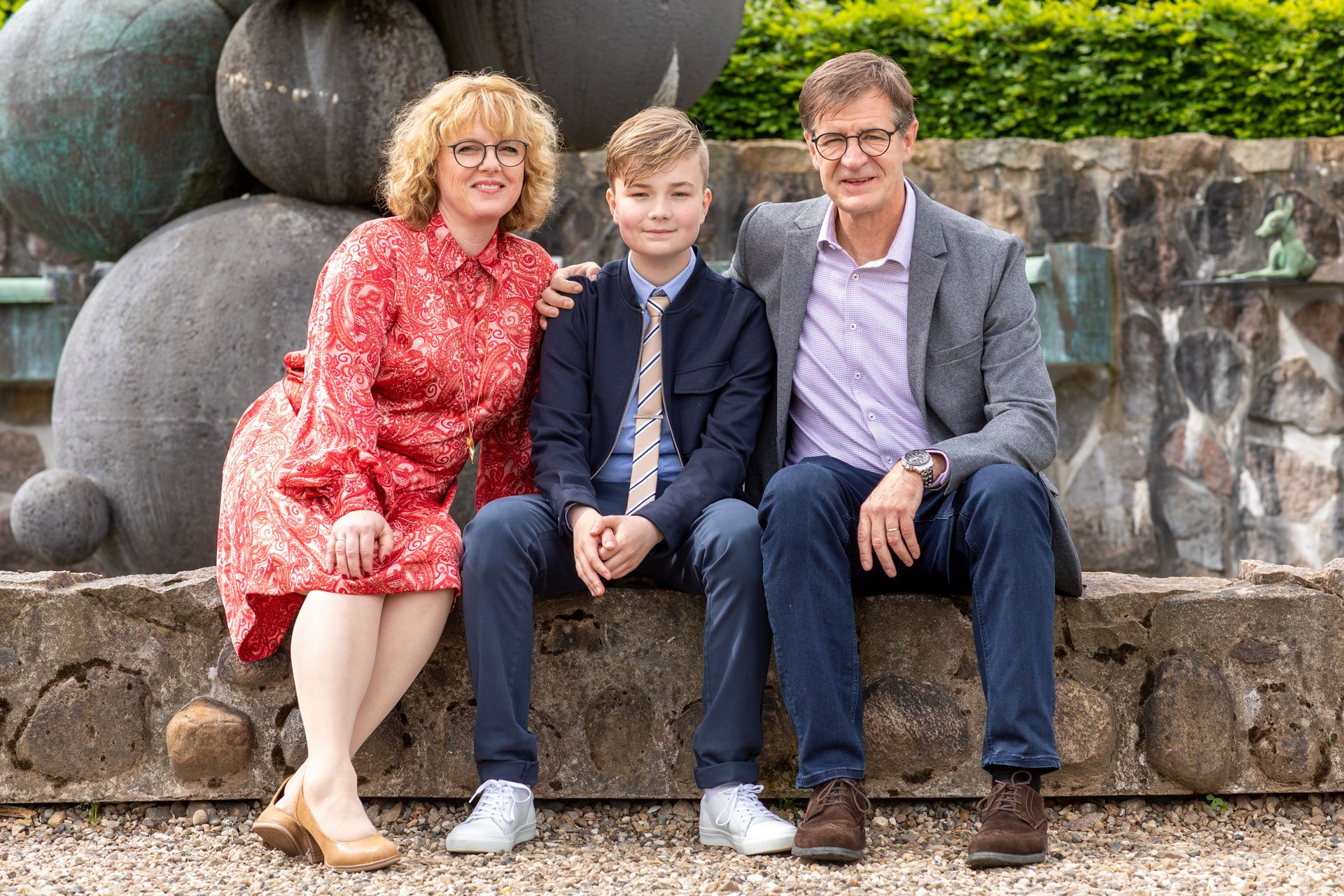 En familie med to voksne og et barn sidder sammen på en stenmur i en have.