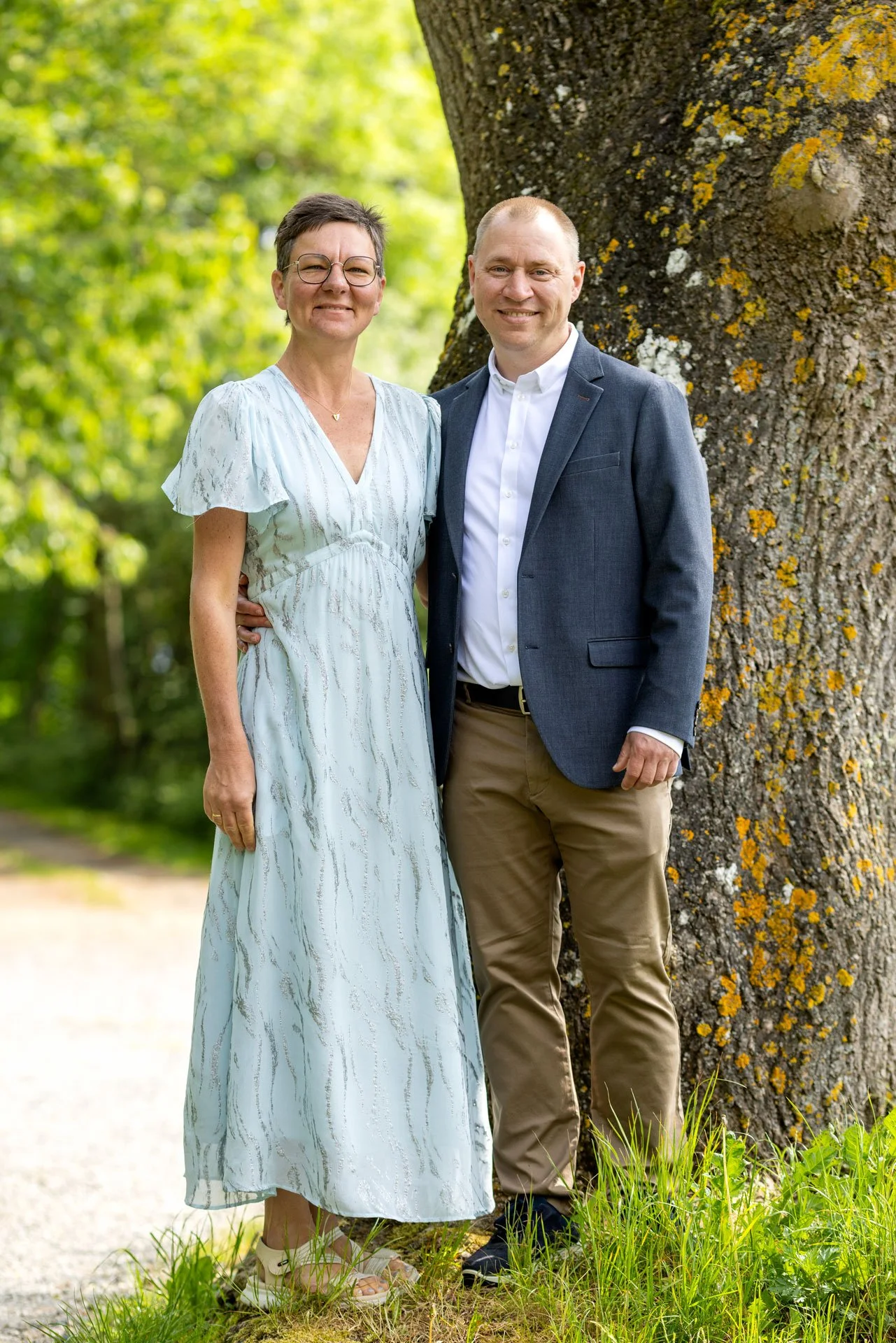 Billedet viser en mand og en kvinde stående ved siden af hinanden foran et stort træ på en græsplæne med grønne blade i baggrunden. De smiler og ser direkte mod kameraet, hun er iført en lang, lys kjole, og han er klædt i en mørk blazer med hvide skj