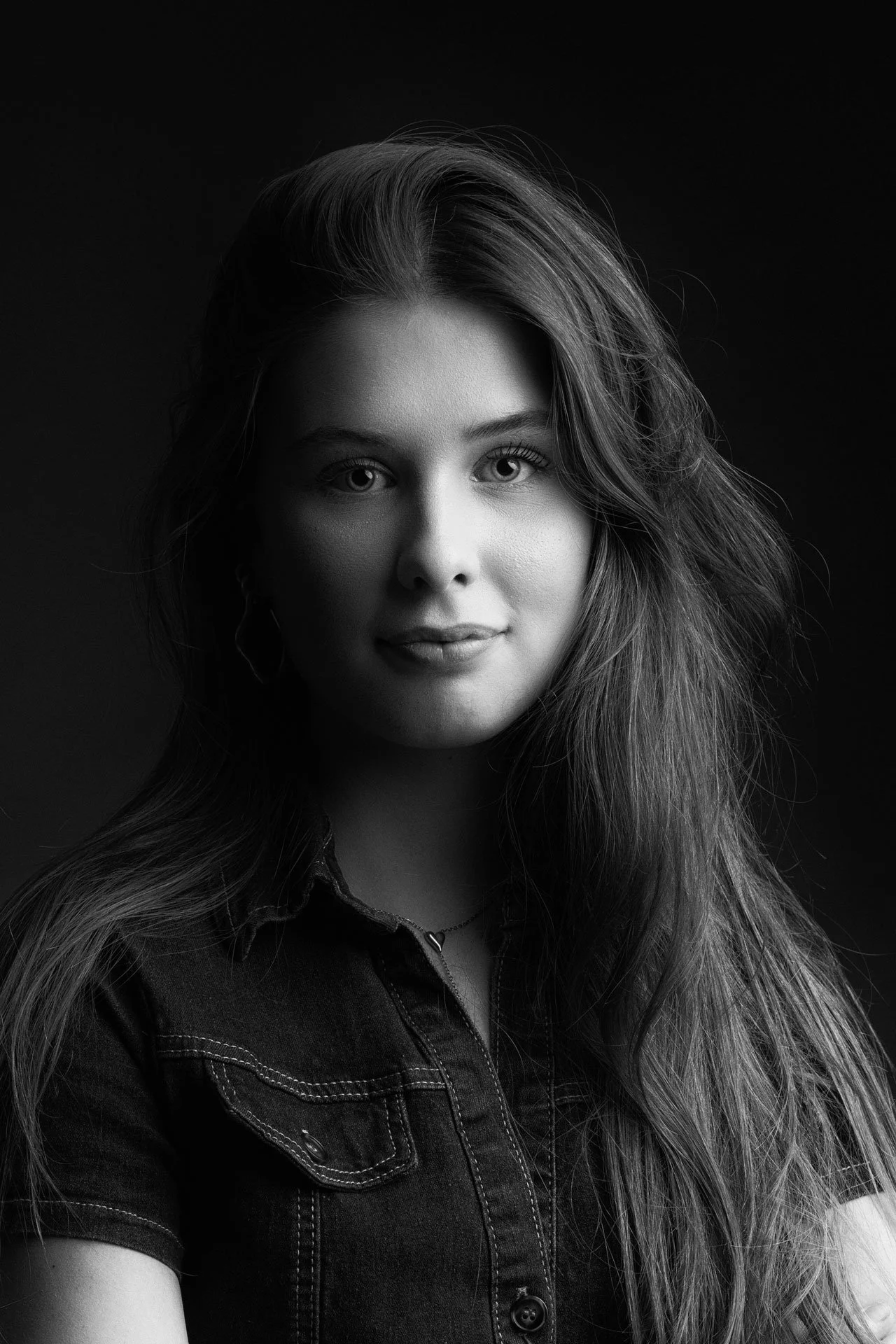 Sort-hvid portrætfotografi af en ung kvinde med langt, vildt hår, iført en denimskjorte, set direkte mod kameraet.