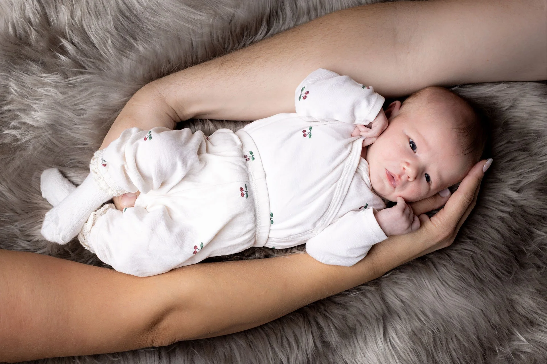 En baby ligger på en blød, grå pelsdyne, holdt forsigtigt af en persons hænder.