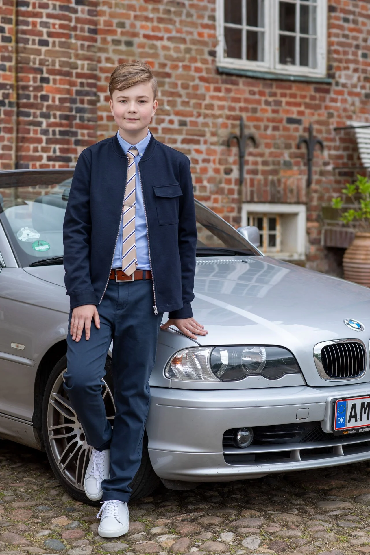 En dreng står ved en sølvgrå BMW bil foran en murstensbygning. Han er iført blå skjorte, blå bukser, en beige og blå stribet slipse og en mørkeblå jakke, og har lysebrunt hår.