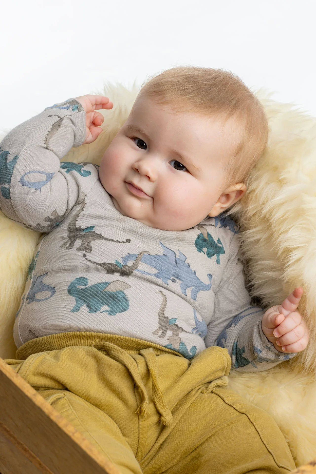 Billedet viser en glad ung dreng med lyst hår, iført en grå T-shirt med dino-mønster og gule bukser, liggende på en blød, varm pels-agtig overflade.