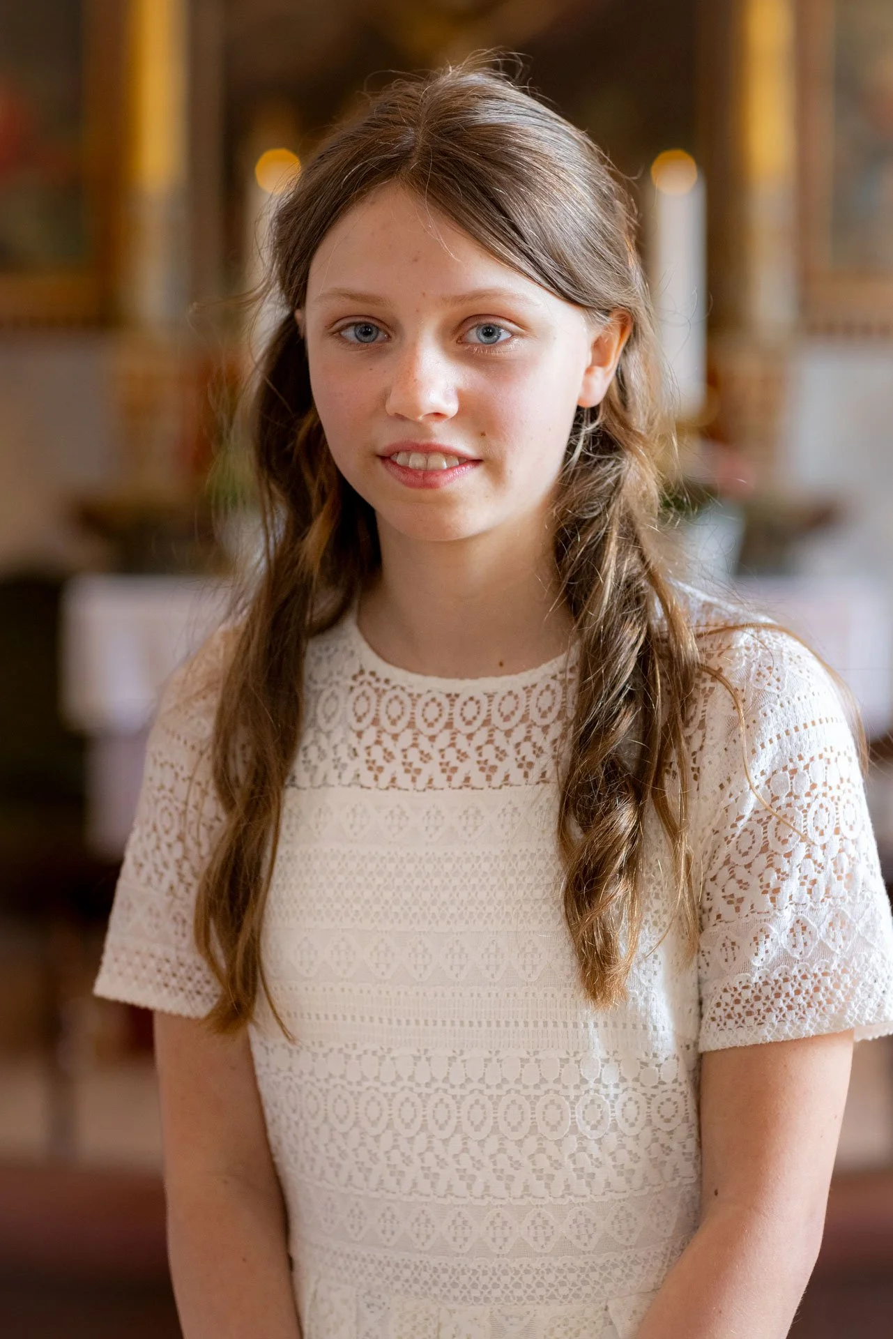 En ung pige med langt, brunt, bølget hår og blå øjne, iført en hvid blondetop med broderi, står indendørs med en sløret baggrund.