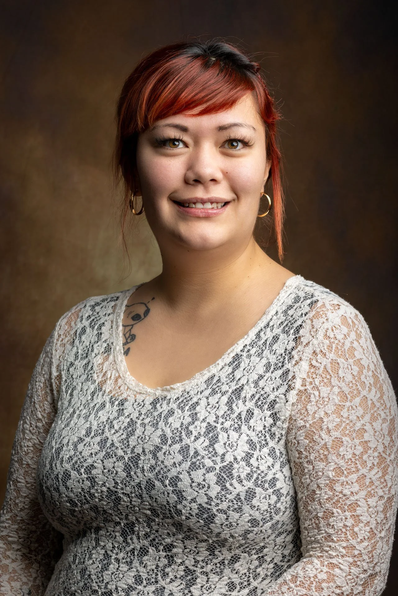 Portræt af en kvinde med rødt hår, guldbøjlerøreringe, og en tatovering på venstre skulder, iført en hvid blondebluse.