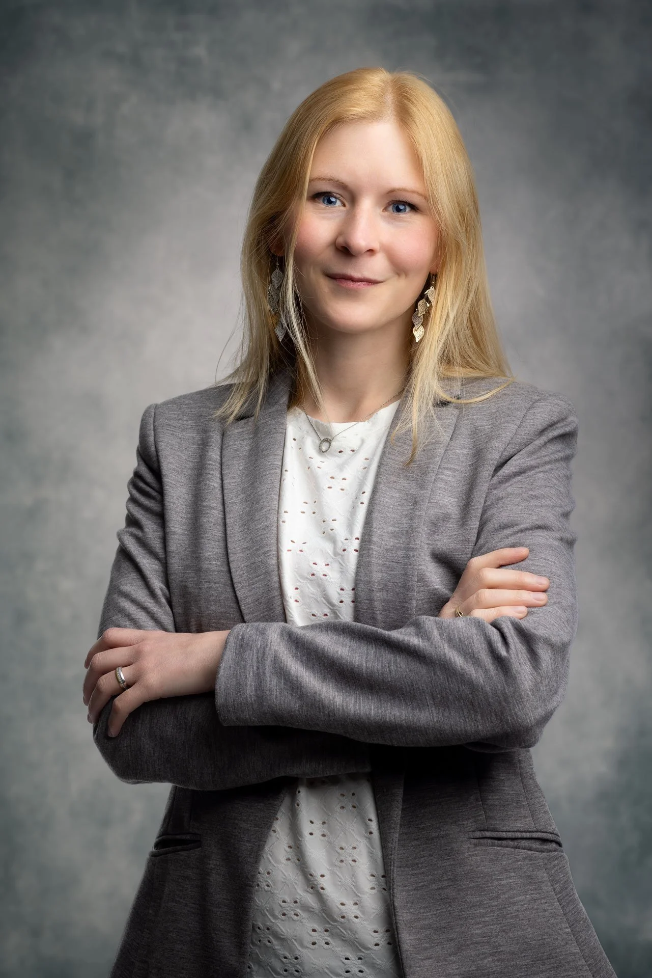 Kvindelig portræt af en mor med blond hår, iført grå blazer og hvid kjole med hulmønster, poserer med armene krydset mod en grå baggrund.