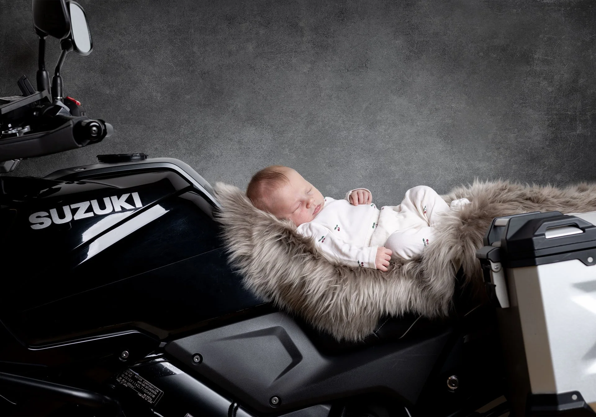 En baby ligger og sover på en faux-fur pude på en sort Suzuki motorcykel, med en grå baggrund.