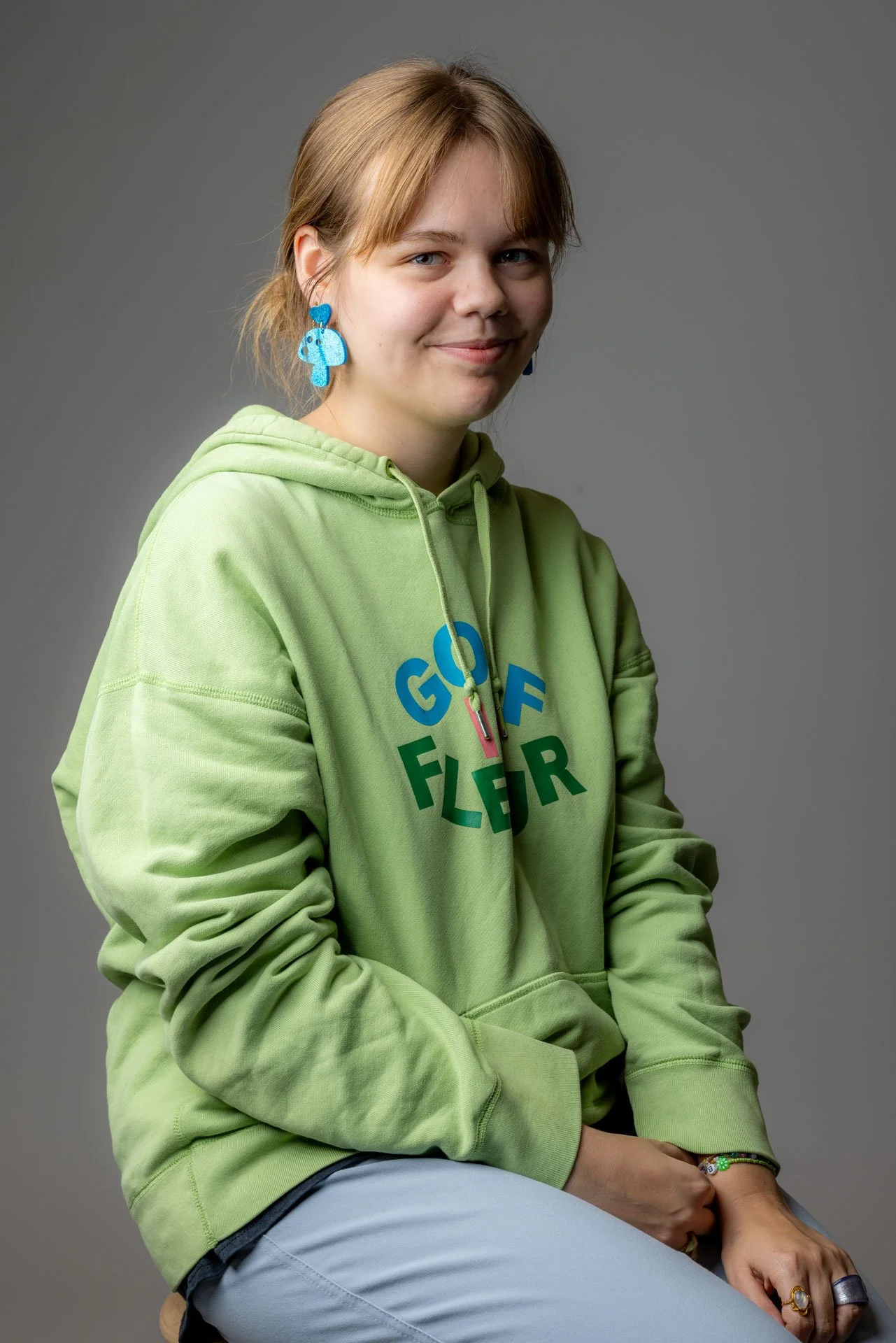 En ung kvinde med lysebrunt hår iført en lysegrøn hættetrøje med teksten 'GOLF FLEJR', blå øreringe og flere ringe, sidder mod en grå baggrund.