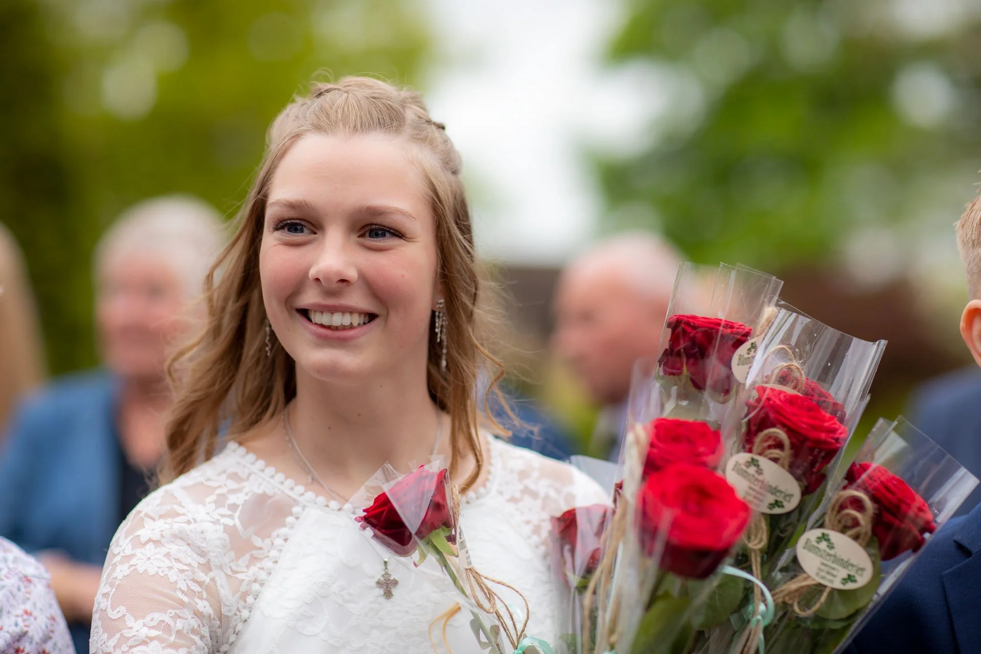 Smilende ung kvinde med krøllet, halvlang hår i hestehale, iført hvid blondet kjole, holder buket røde roser, omgivet af mennesker, udendørs med grønt træ i baggrunden.