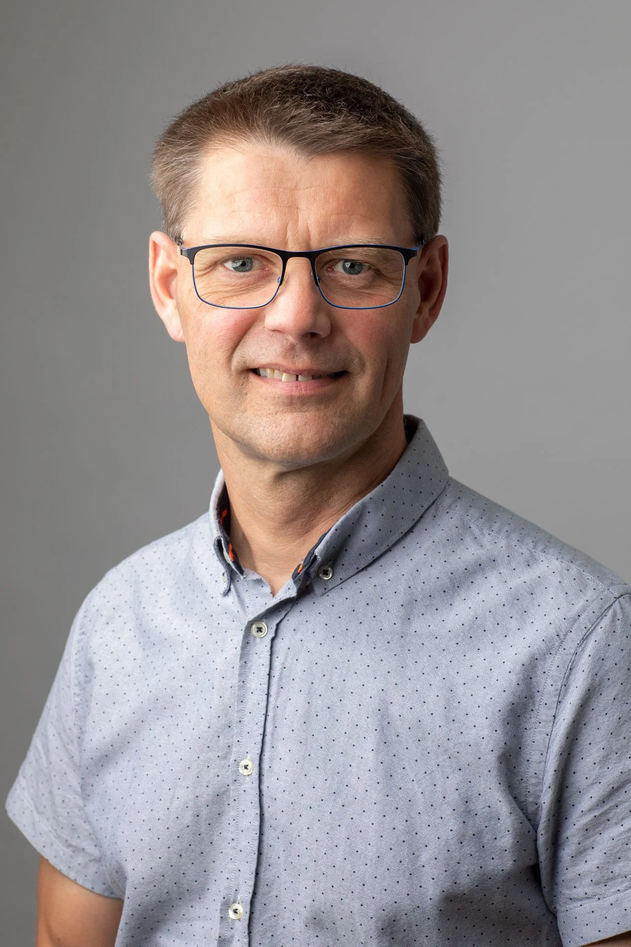 Portræt af en mand med briller, smilende, iført en lyseblå skjorte med små polkaprikker, mod en neutral grå baggrund.