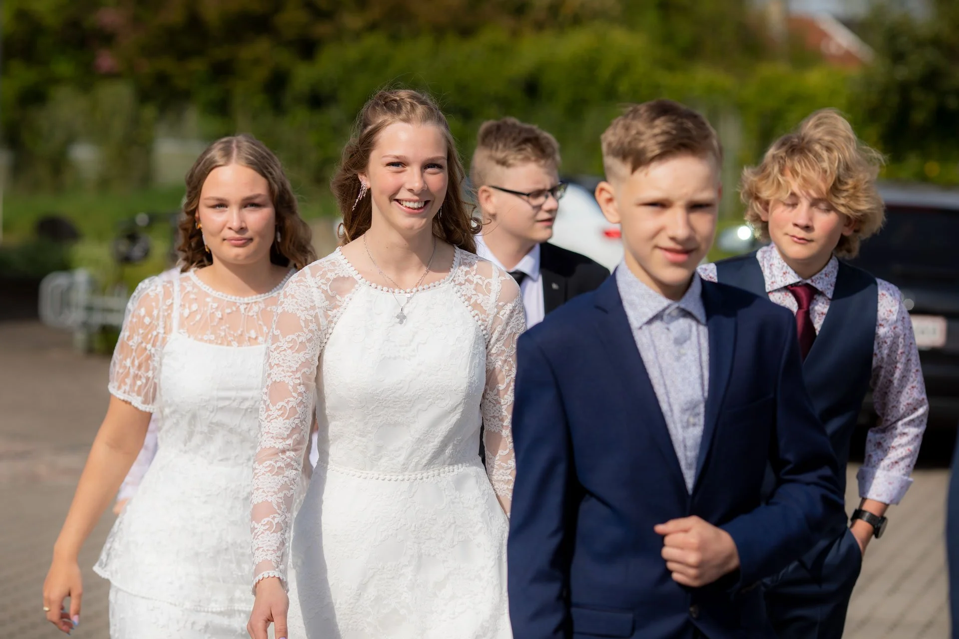 En gruppe unge mennesker går sammen ude i det grønne, nogle er klædt i festlige kjoler og jakker.