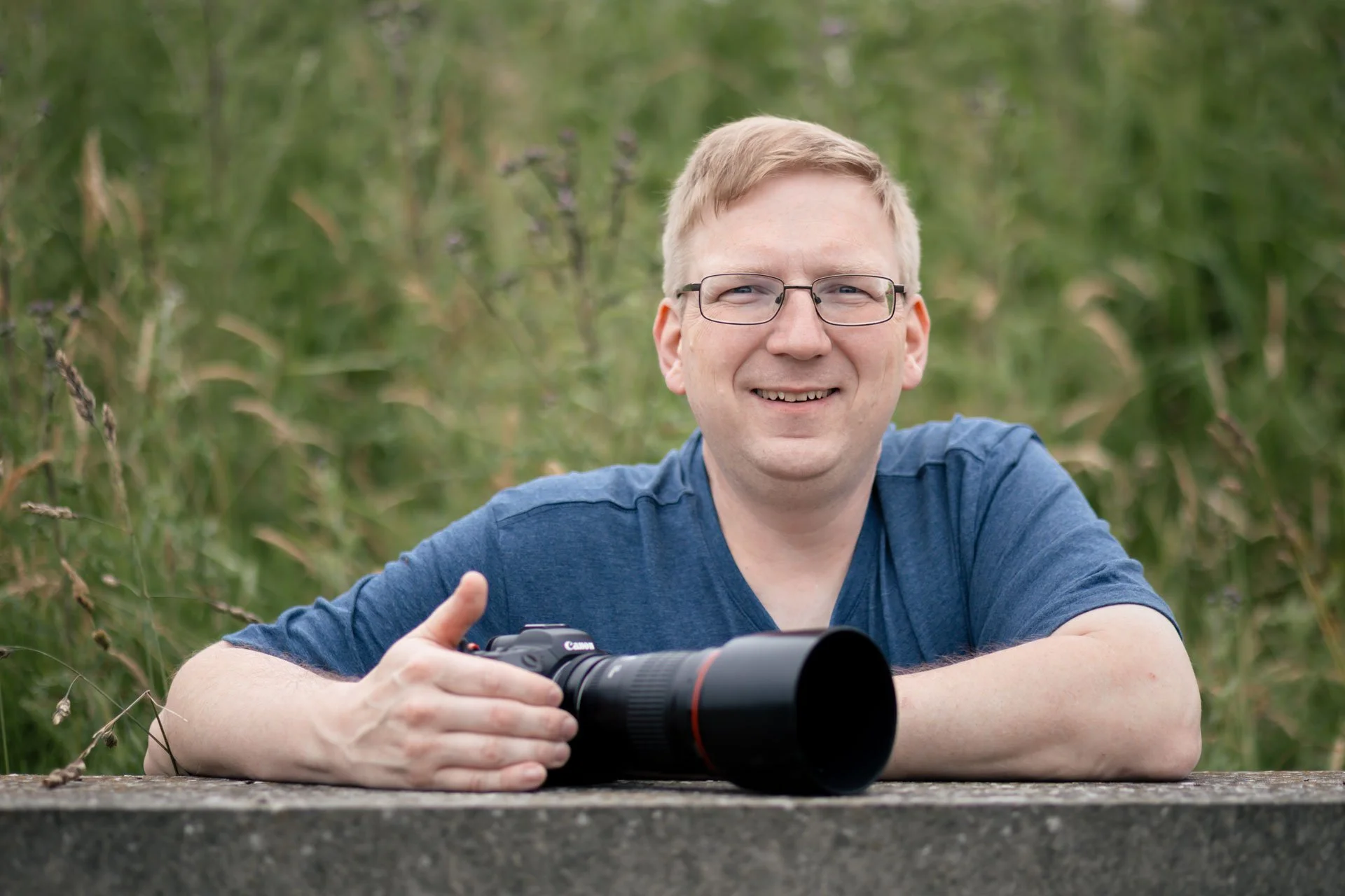 Mand med briller, smilende, holder et teleobjektiv kamera, sidder ved en sten, grøn vegetation i baggrunden.