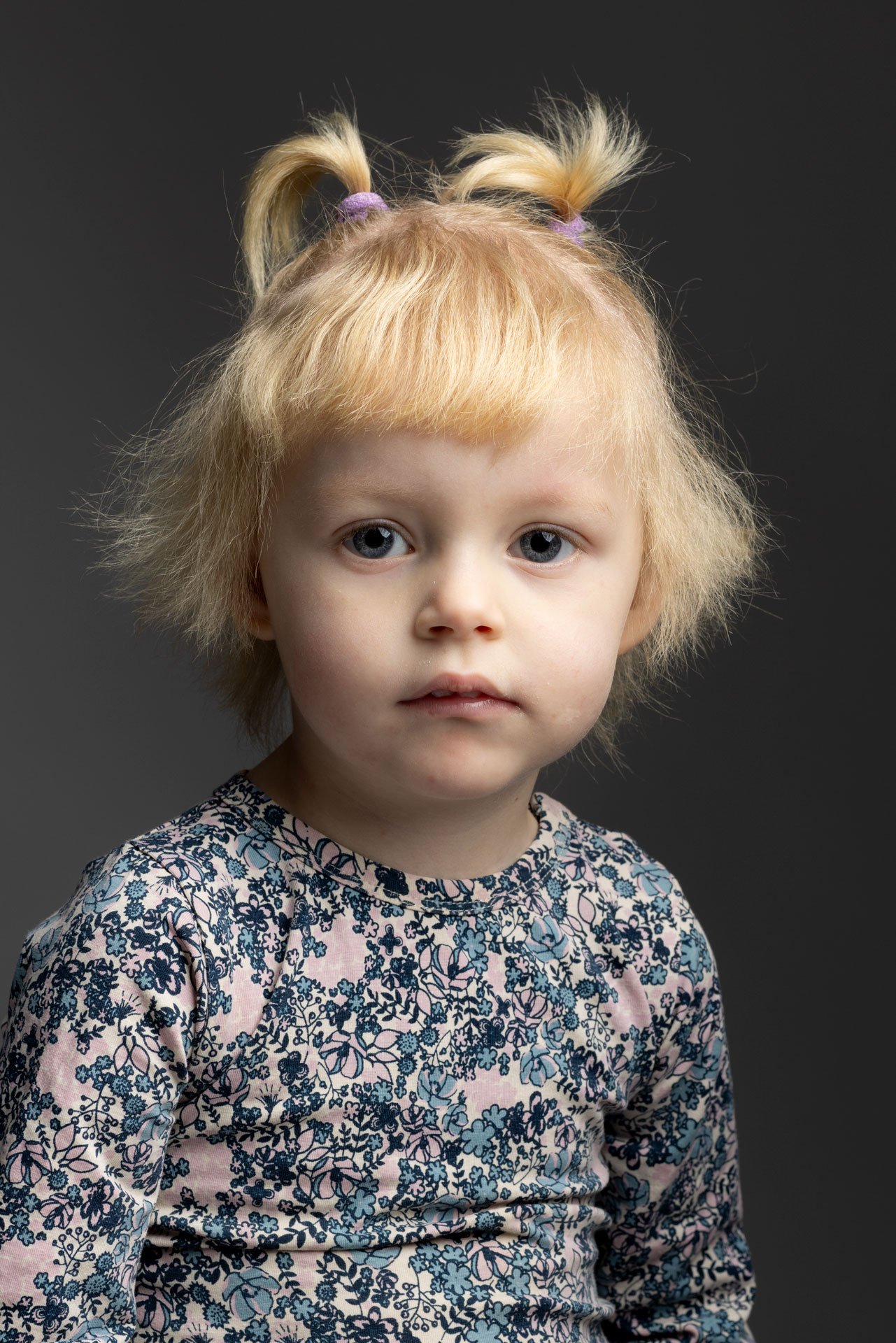 Portræt af en ung pige med lysekrøllet hår i to tovs, iført en blomstret langærmet bluse, mod en mørk baggrund.