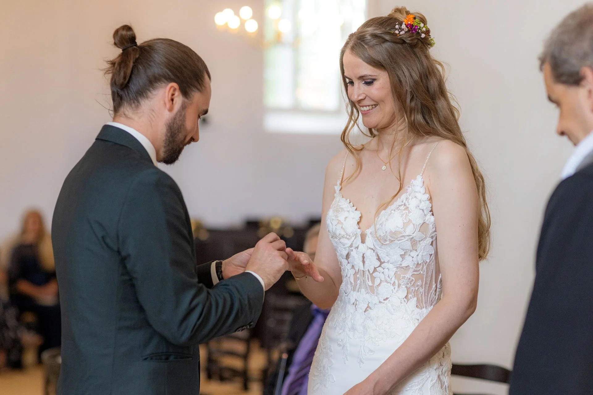 Et bryllupsceremonie med en kvinde i en hvid brudekjole og en mand, der giver hende et forlovelses- eller vielsesring, mens en anden mand står ved siden af.