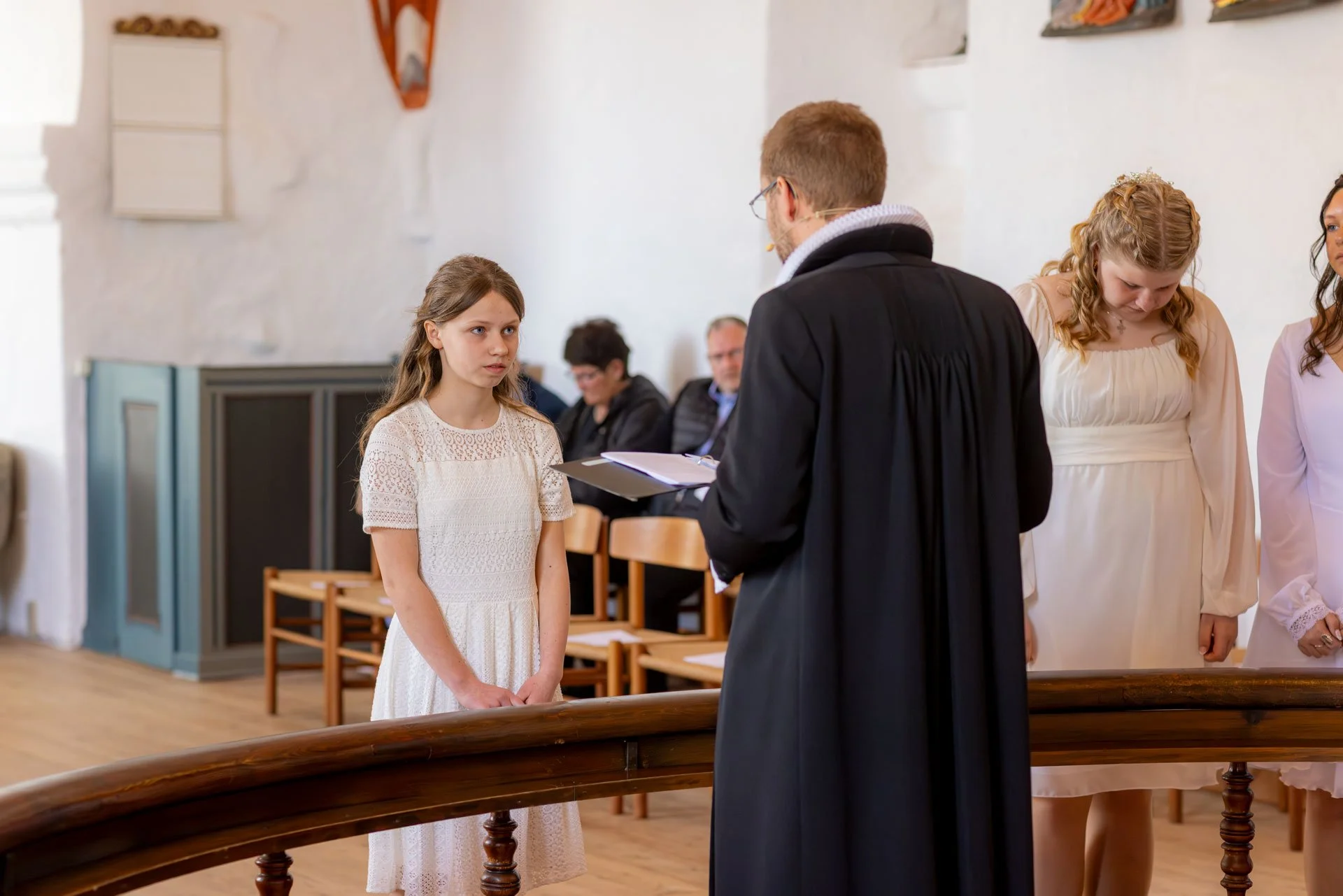 En pige står foran en præst under en ceremoni, sandsynligvis en konfirmation, i en kirkesal med andre tilskuere i baggrunden.