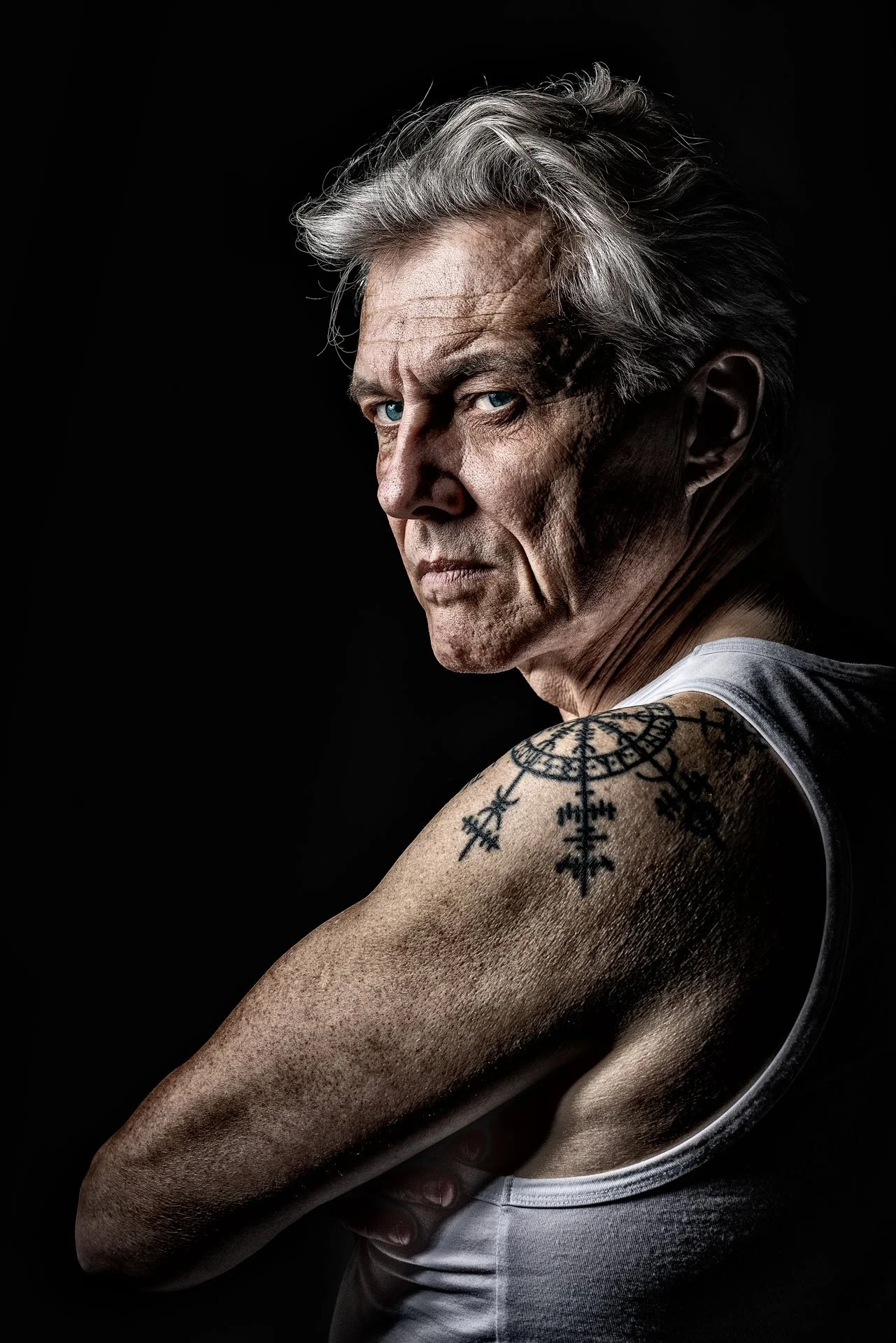 Et portræt af en ældre mand med gråt hår, iført en hvid tanktop, der viser en tatovering på armen, set mod en sort baggrund.