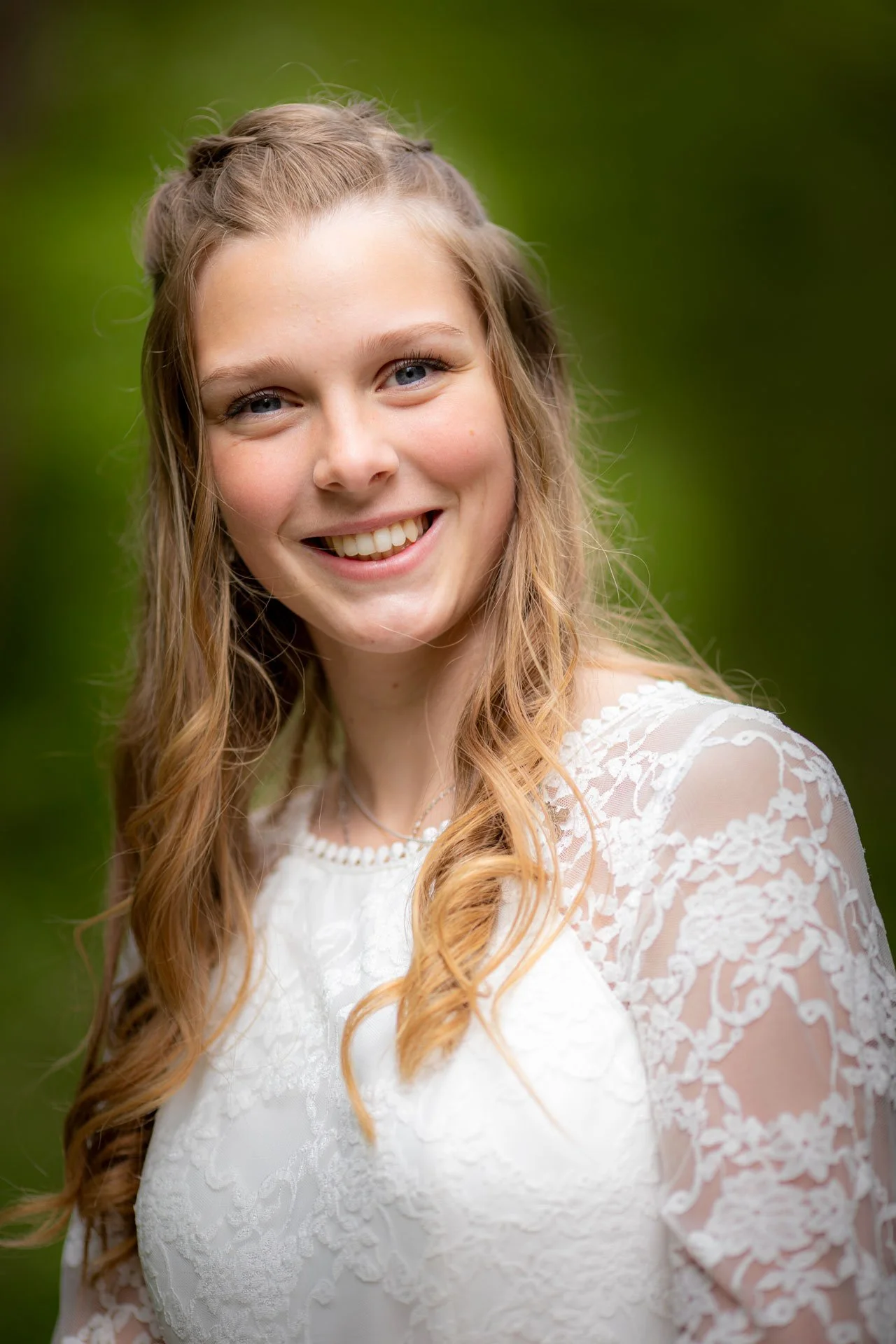 Ung kvinde med langt, bølget, blond hår iført en hvid blød kjole med blonder, smiler og ser mod kamera, baggrund med grønne træer.