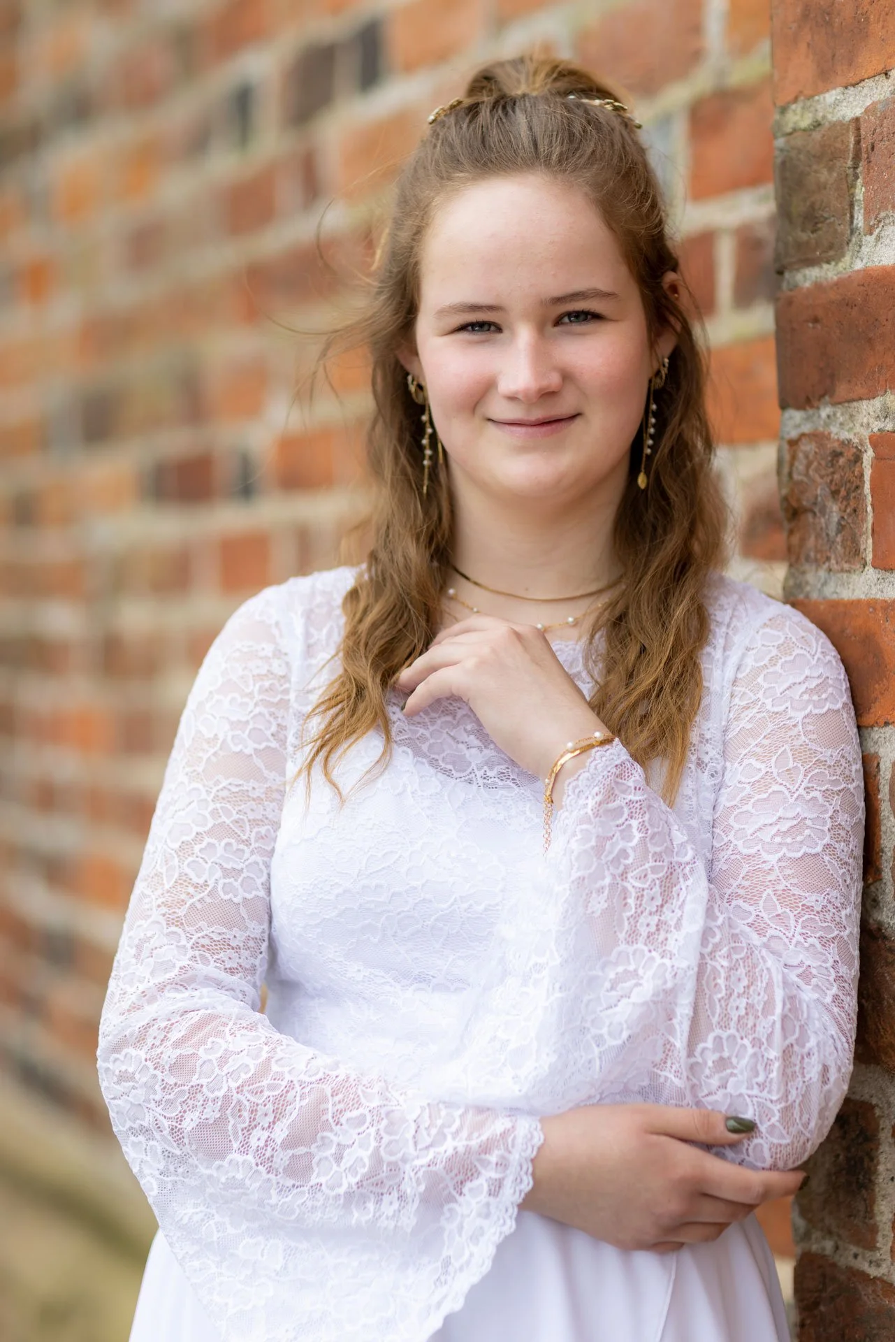 En ung kvinde med langt, krøllet hår, iført en hvid blondes kollet kjole og smykker, står ved en mur og smiler til kameraet.