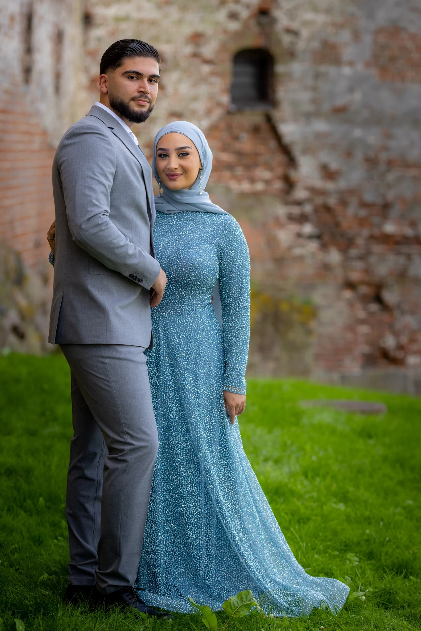 Et par står på en grøn plæne foran en gammel murstensvæg, klædt i formelle tøj, kvinden i en blå kjole med glimmer og blå tørklæde, og manden i en grå dragt.