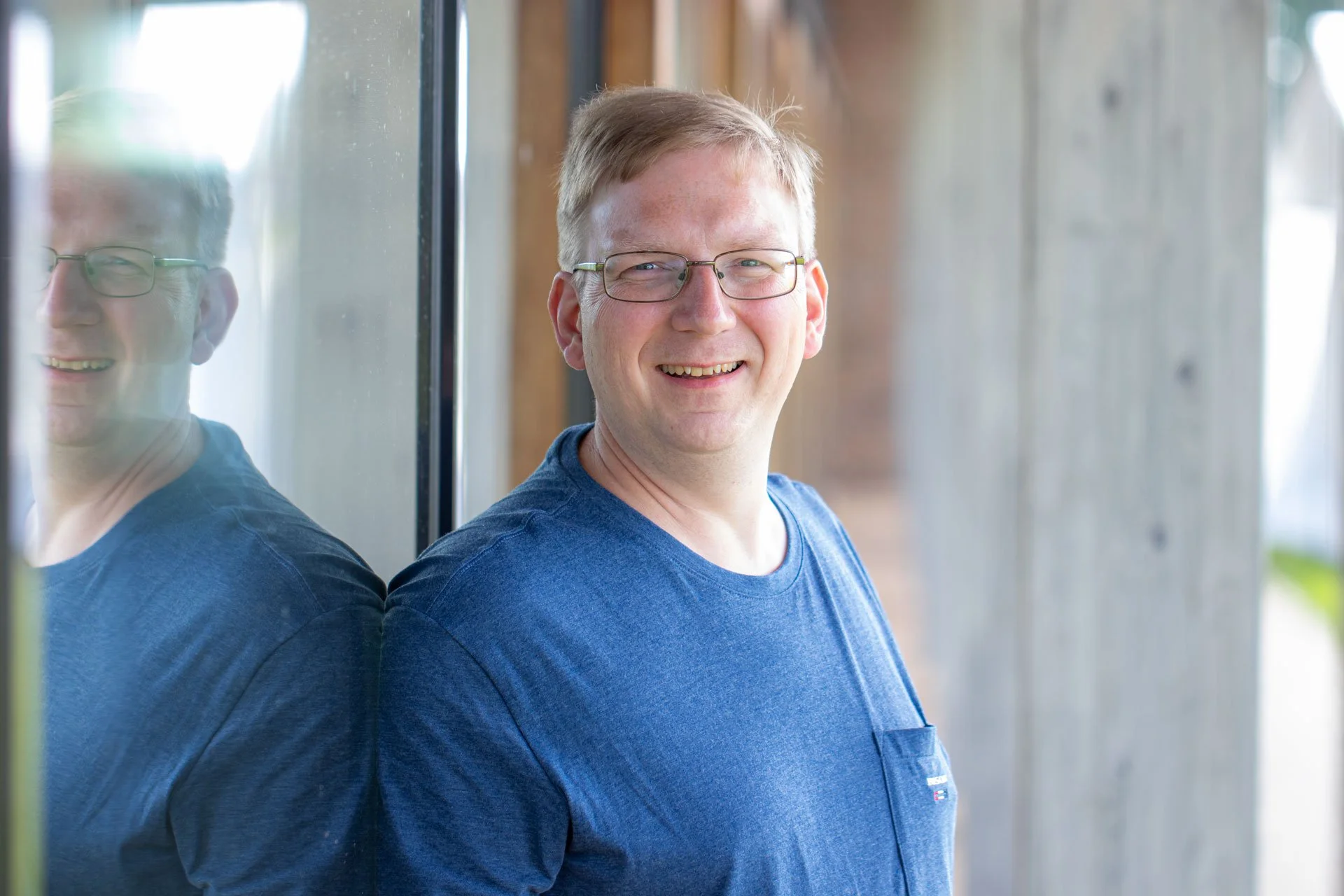 Et smilende mand med briller står ved en væg, med refleksion i ruden.