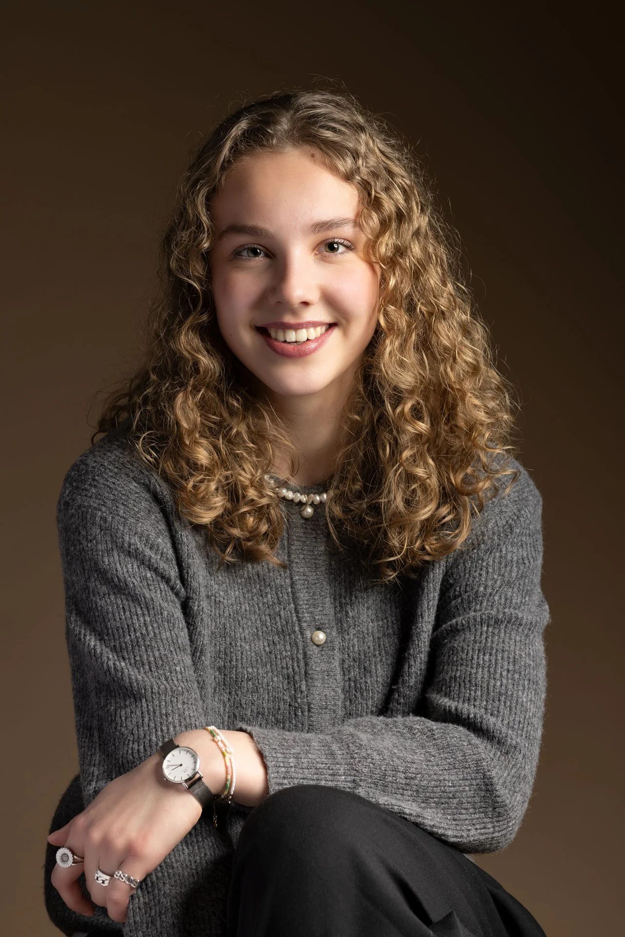 Portræt af en smilende ung kvinde med krøllet hår, iført gråt tøj og smykker, siddende mod en brun baggrund.
