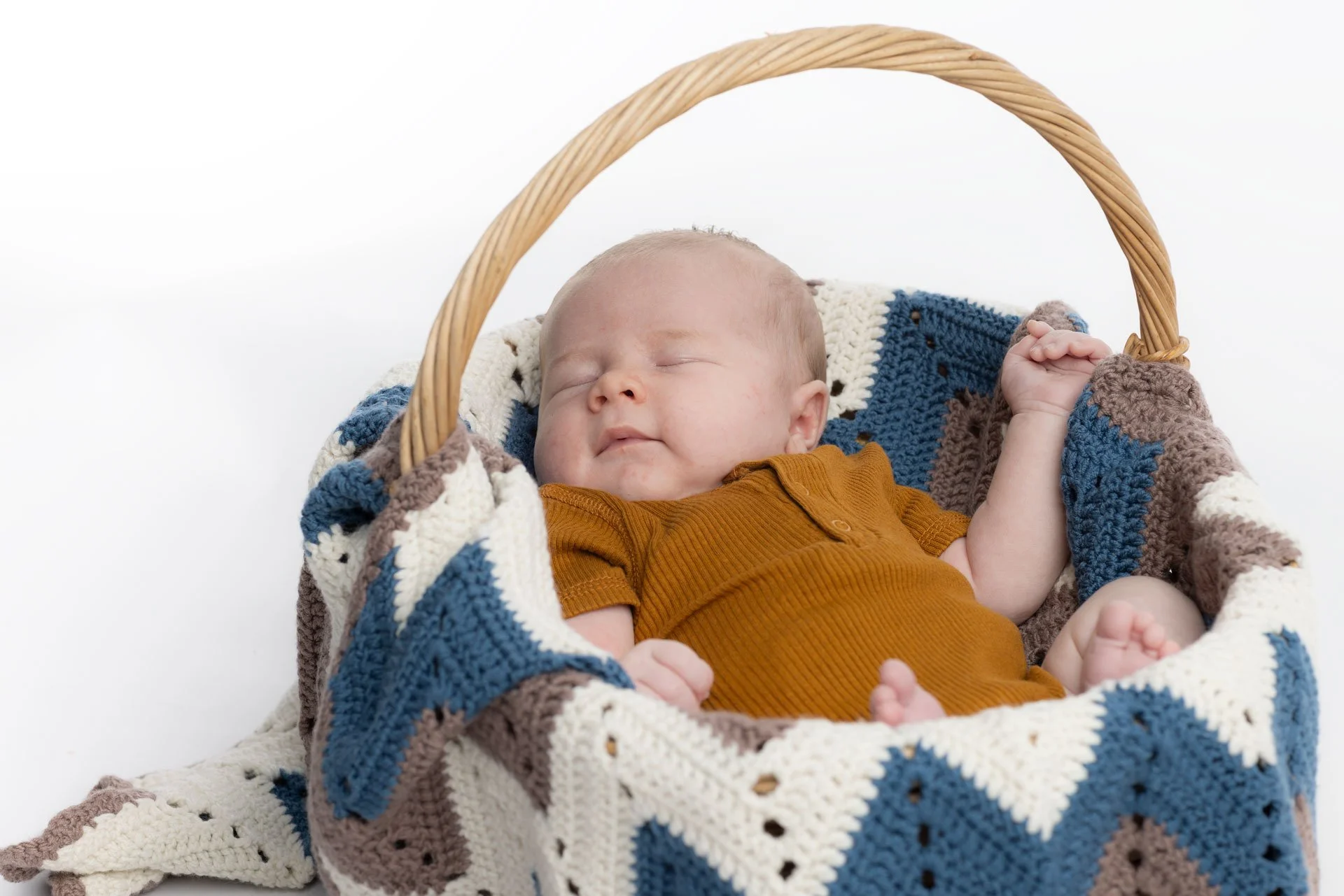 En sovende baby i en kurv, omgivet af en håndstrikket patchwork-tæppe i farverne creme, blå, brun og grå, iført en brun body.