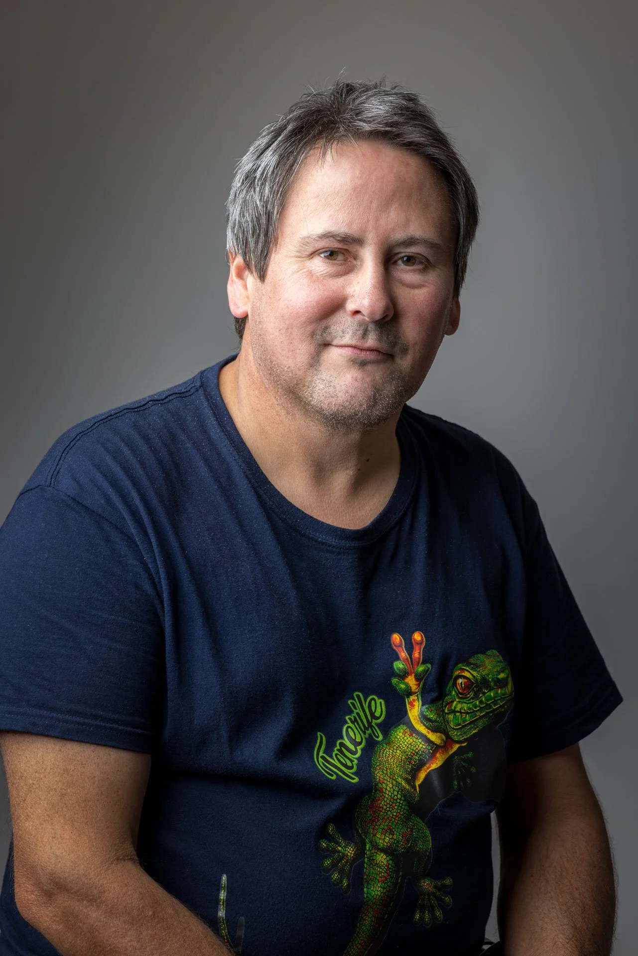 En mand med gråt hår iført en sort T-shirt med en farverig kameleon på.