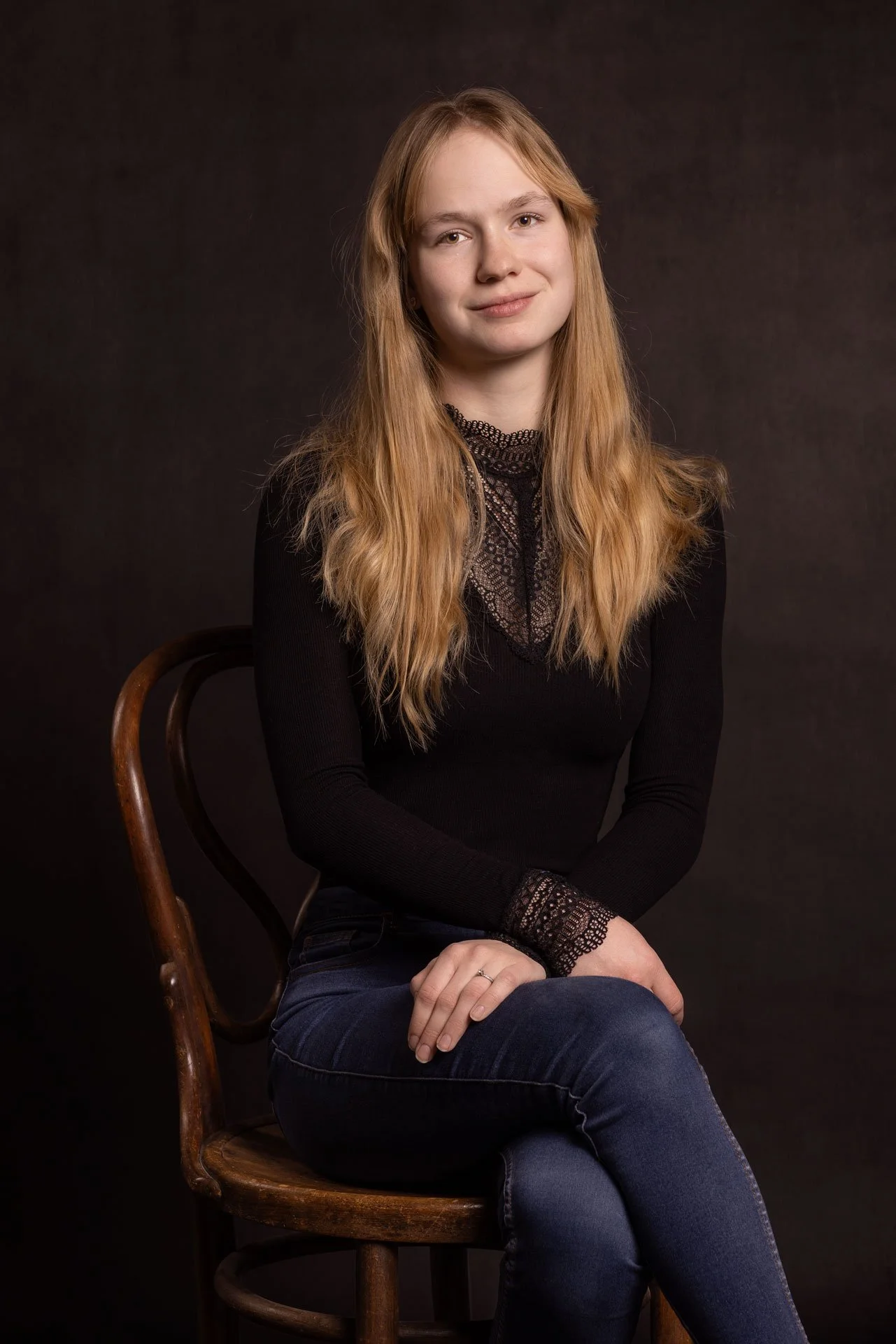En ung kvinde med langt, lyst hår sidder på en træstol mod en mørk baggrund. Hun har på en sort, langærmet top med blonde detaljer ved hals og håndled, og hun smiler let.