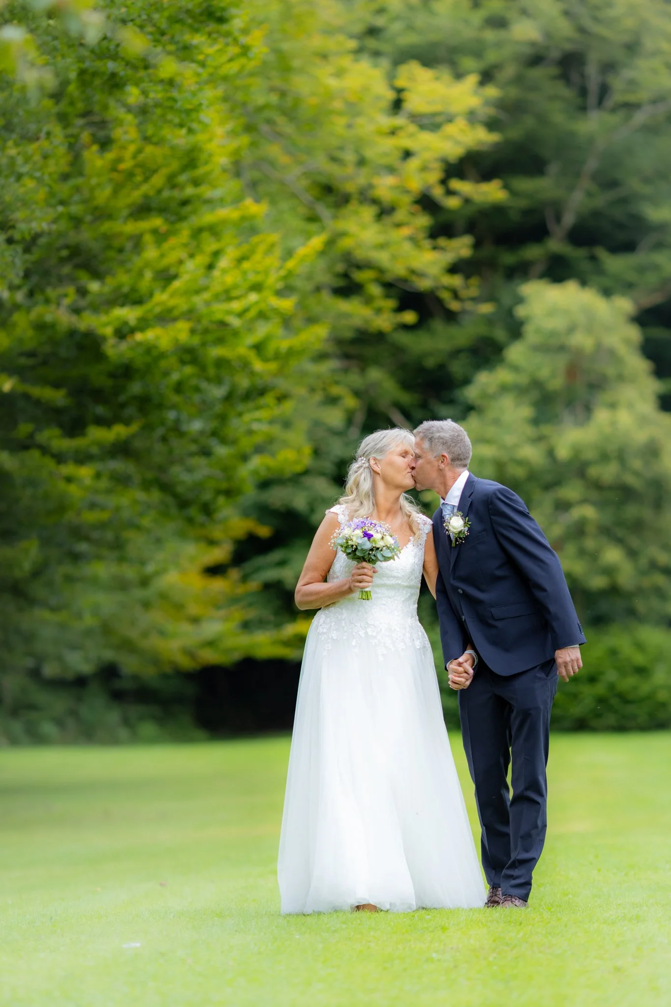 Et ægtepar kysser på en grøn græsplæne foran grønne træer, hvor kvinden bærer en hvid brudekjole og holder en blomsterbuket, og manden er i en mørk blå jakkesæt.
