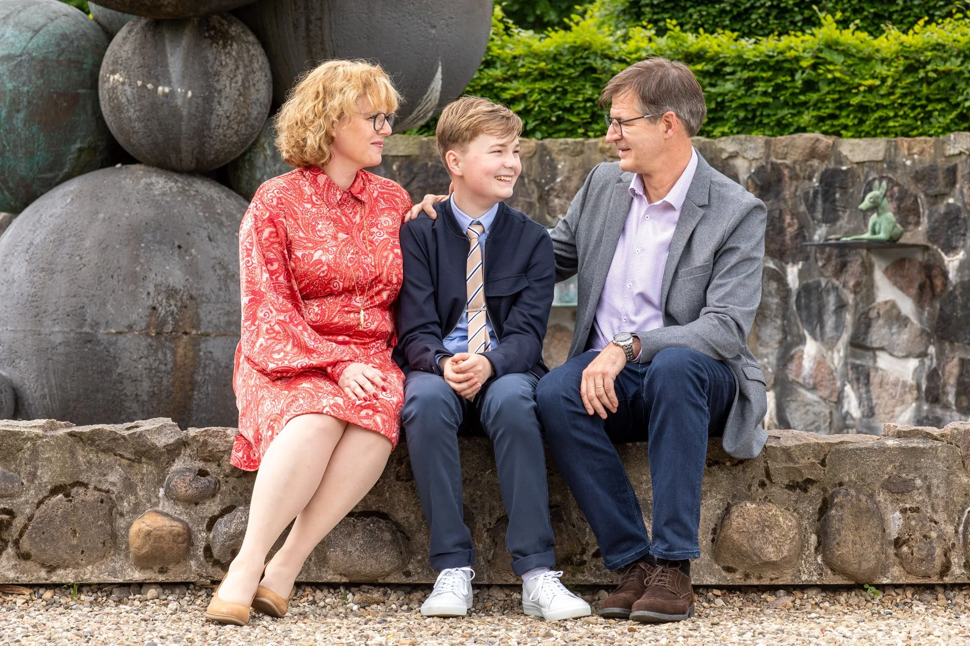 Tre personer sidder på en stenmur udendørs. Der er en voksenkvinde med lyst krøllet hår i en rød kjole, en dreng i skoleuniform og en voksen mand i en grå blazer, der smiler og ser på hinanden.