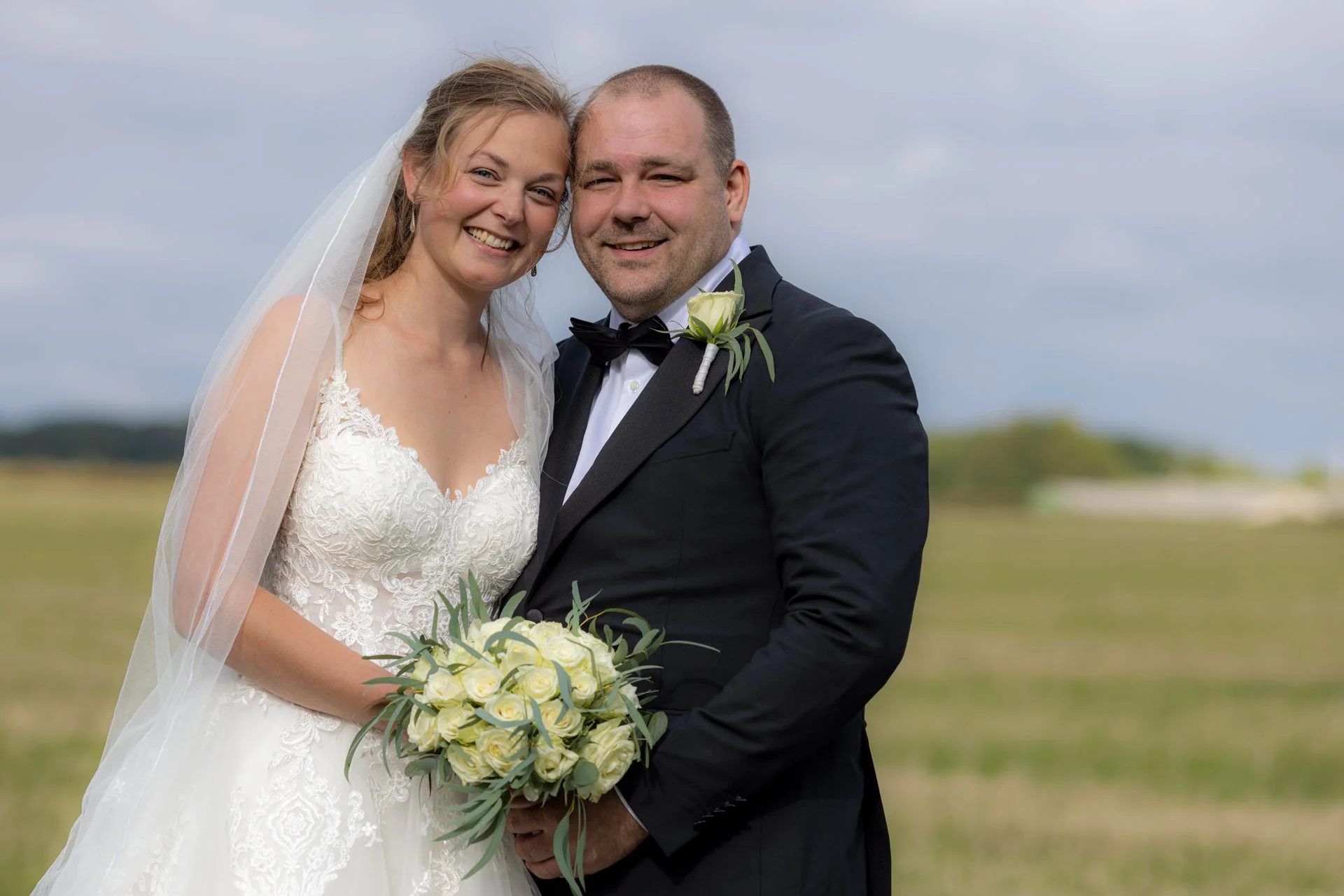 En glad brud og brudgom står sammen udenfor i et åbent landskab, iført festkjole og smoking.