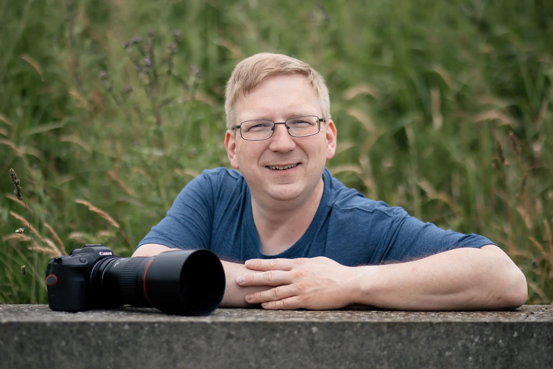 Mand sidder på en concrete bund med et kamera foran sig, omgivet af grønne planter og græs.