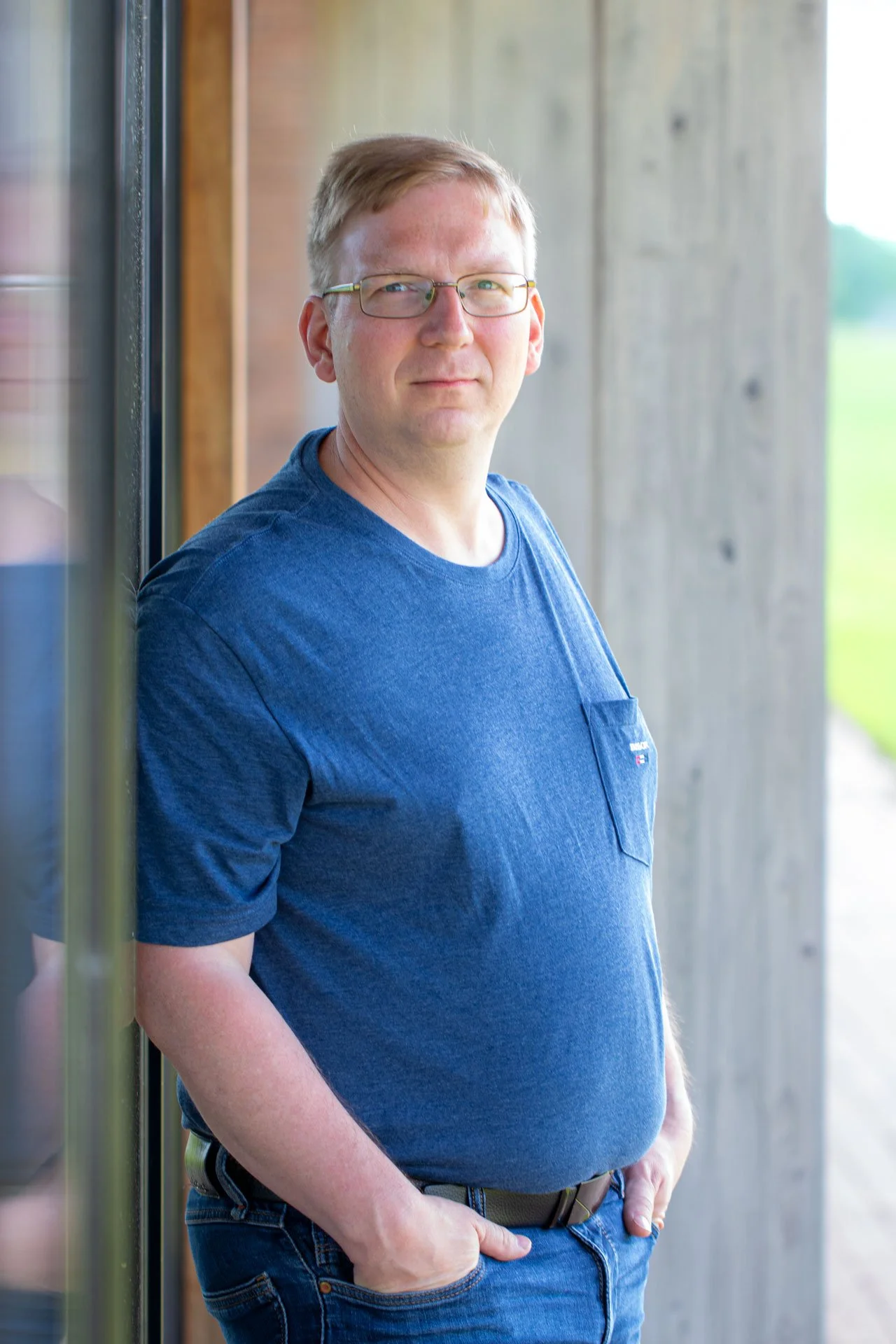 Mand står udendørs ved en trævæg, iført blå t-shirt og briller, og ser roligt mod kameraet.