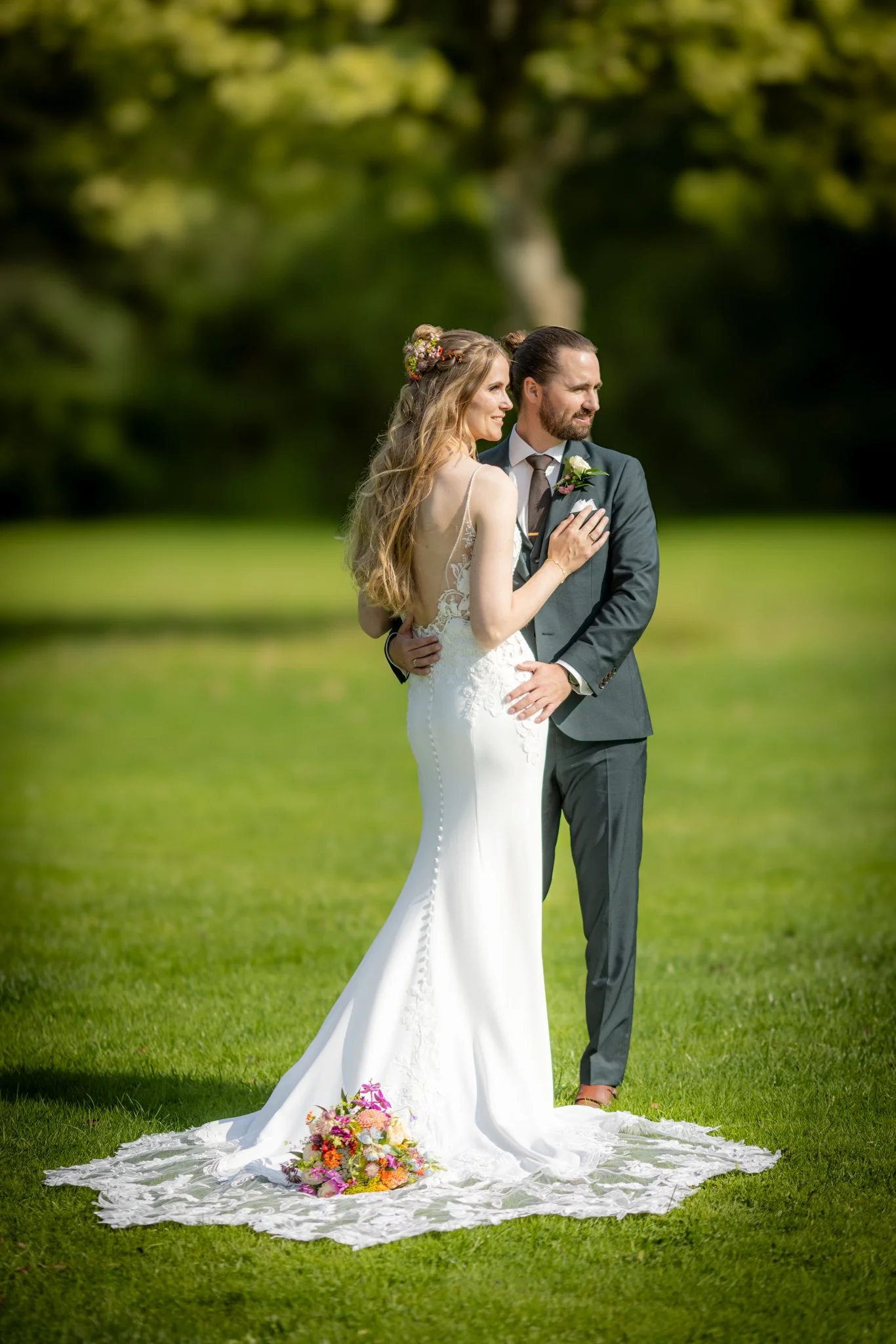 Et brudepar står sammen på en grøn mark, omgivet af træer i baggrunden. Bruden bærer en lang, hvid brudekjole med snørresløjfe på ryggen og en blomsterkrans i håret. Brudgommen er iført en mørkegrøn habit og slips. De smiler og ser på hinanden, mens en blomsterbuket ligger på jorden ved deres fødder.