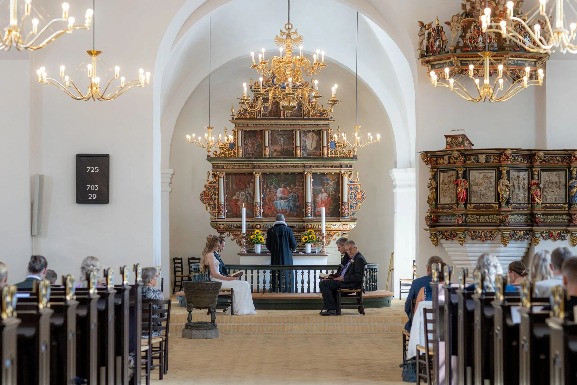 Indendørs billede af en kirke med en præst foran en alter, hvor der er unge mennesker siddende under en ceremoni, omgivet af kråbelysning og gamle religiøse kunstværker.