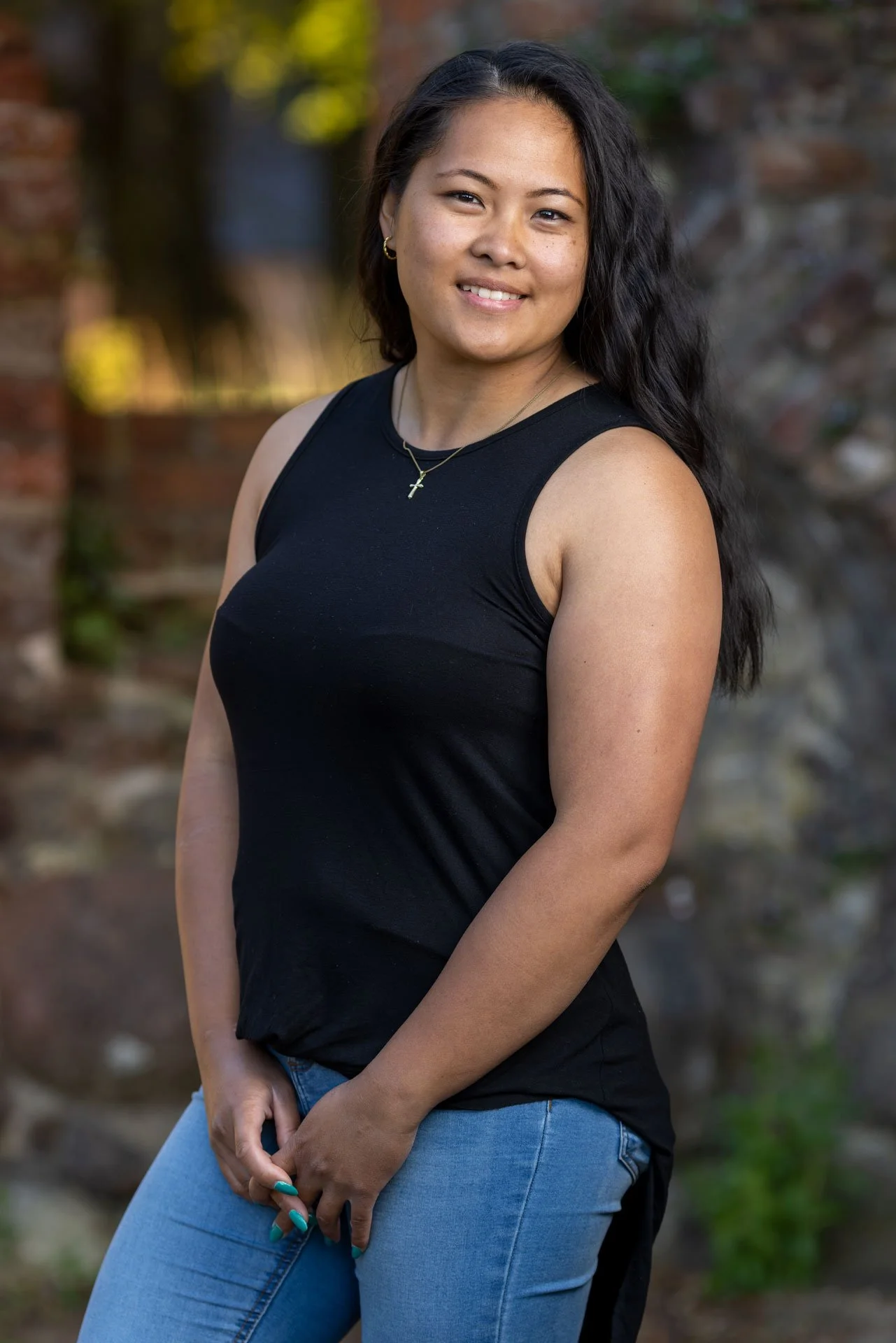 En smilende kvinde med lang sort hår, iført sort top og blå jeans, står foran en mur med en grønt og brunt baggrund.