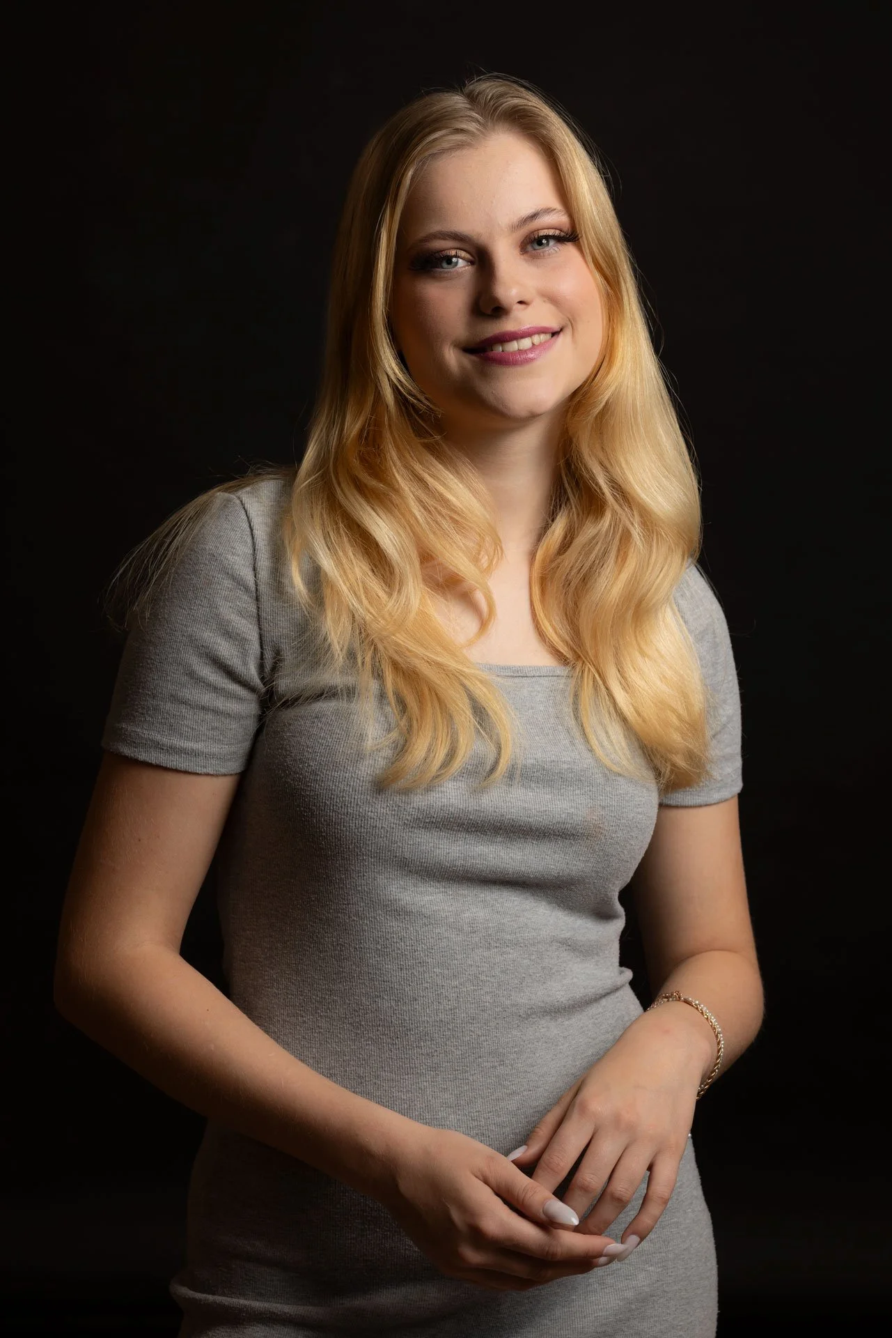 Et portræt af en smilende ung kvinde med langt, blond hår, klædt i en grå t-shirt, mod en sort baggrund.