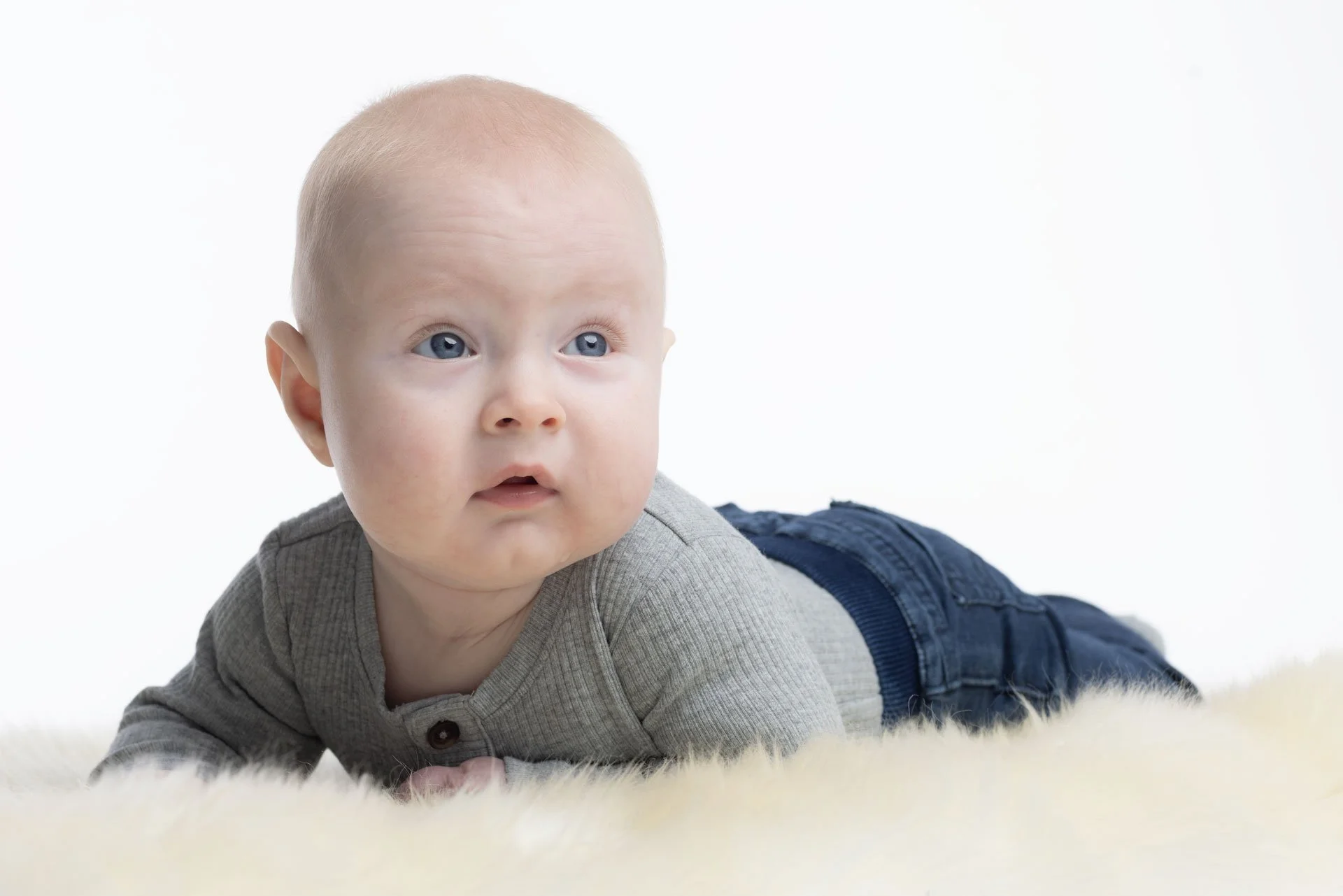 En baby med blond hår og blå øjne ligger på maven på en cremefarvet overflade, iført grå trøje og blå bukser, med en neutral baggrund.