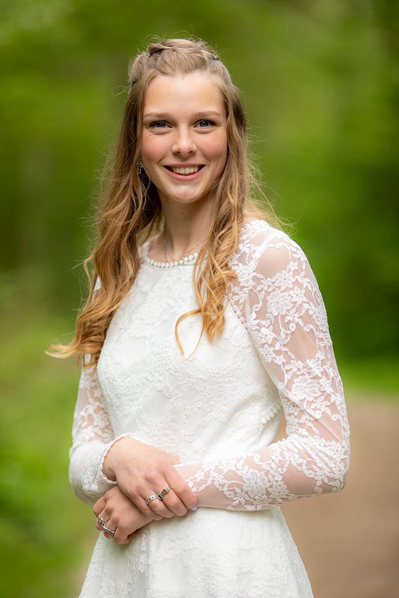 En glad ung kvinde i hvid brudekjole med blondeærmer, stående udendørs med grøn baggrund.