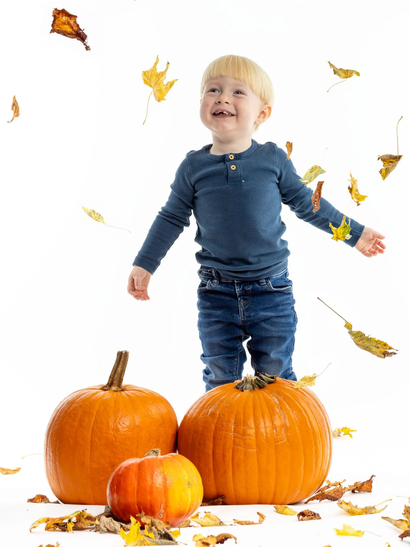 En dreng står mellem tre græskar, mens efterårsblade flyver rundt om ham.