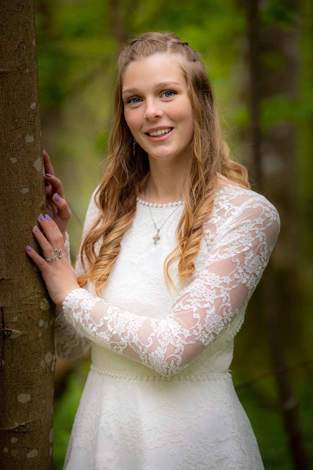 En ung kvinde står udendørs bag en træstam, iført en hvid kjole med blondelook og har lyst hår. Baggrunden er grøn med træer.