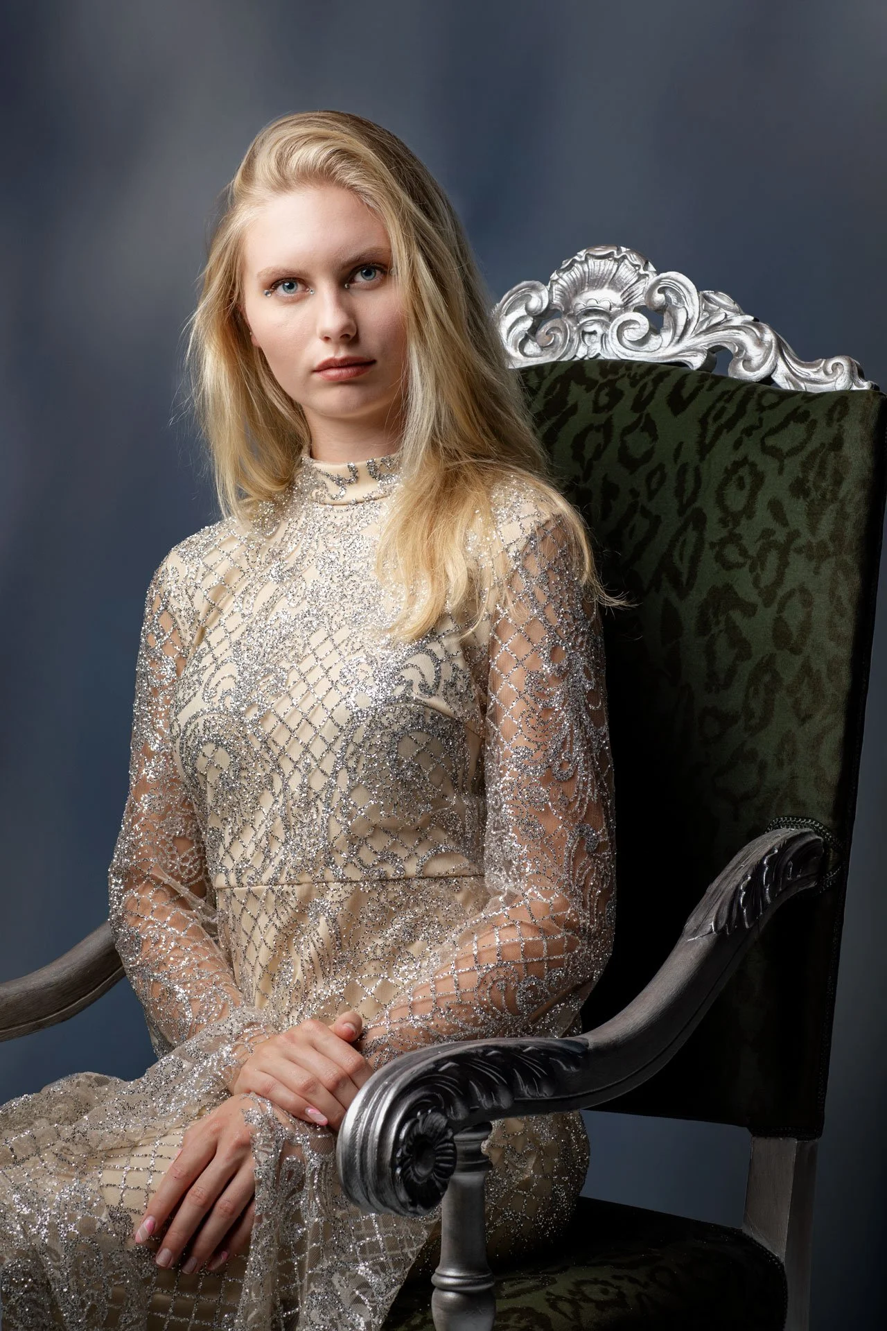 En ung kvinde med langt blondt hår, siddende i en sort og grøn velourstol med detaljer i sølv. Hun bærer en rigeligt dekoreret, metallic-farvet kjole.