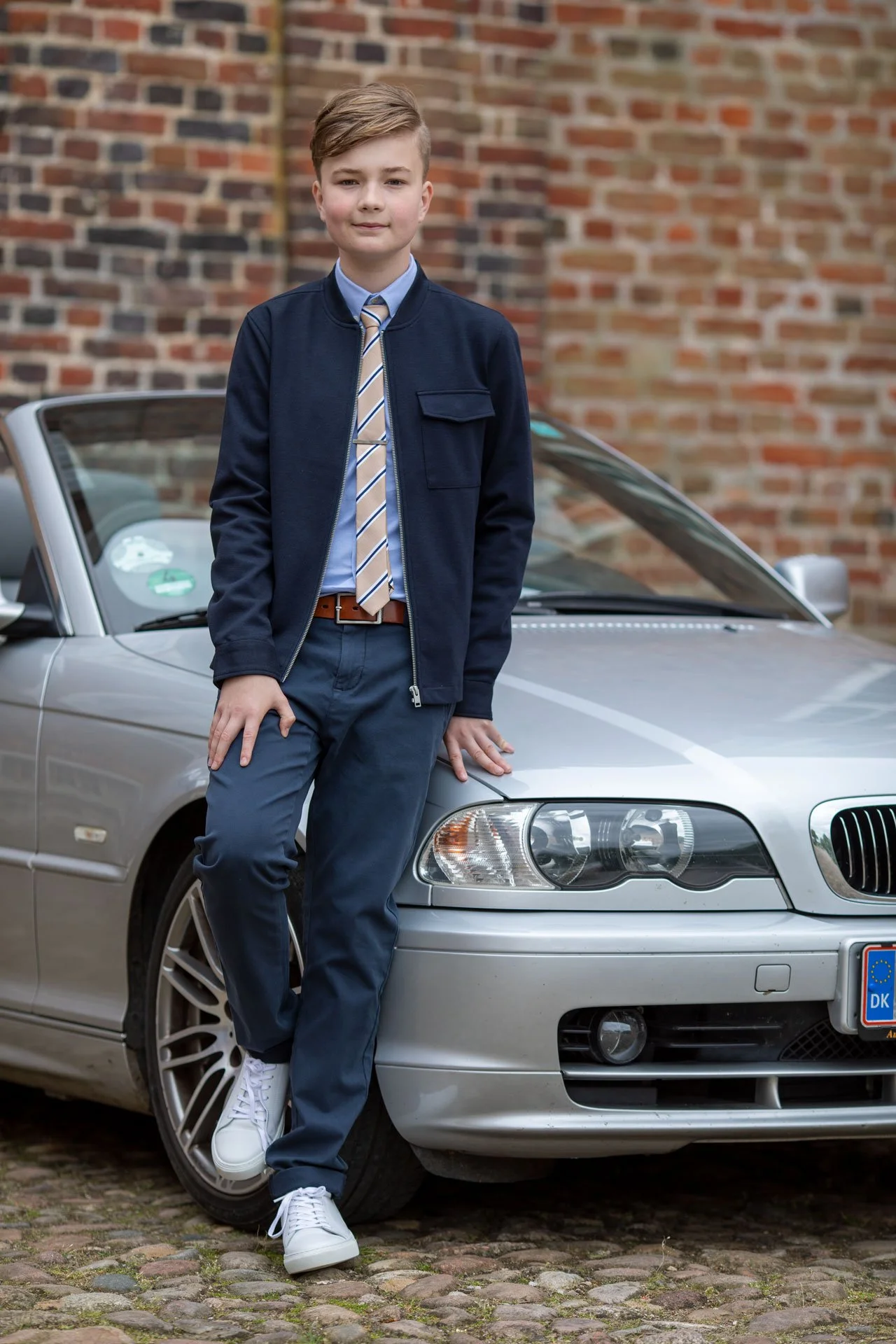 En dreng står ved en sølvfarvet bil på en brostenbelagt vej, foran en gammel murstensvæg. Han har kort, lyst hår og bærer en blå jakke, en lys blå skjorte, en ternet slips, mørkeblå bukser og hvide sneakers.