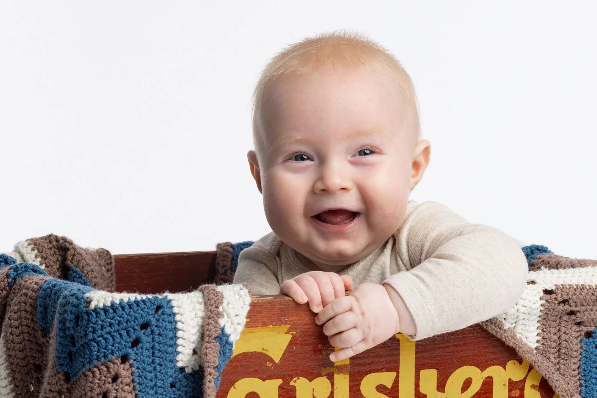 Et smilende baby med blond hår, der holder fast i kanten af en farvet trækasse, der er dækket med en håndstrikket tæppe. Babyen har på sig en lys beige langærmet trøje.