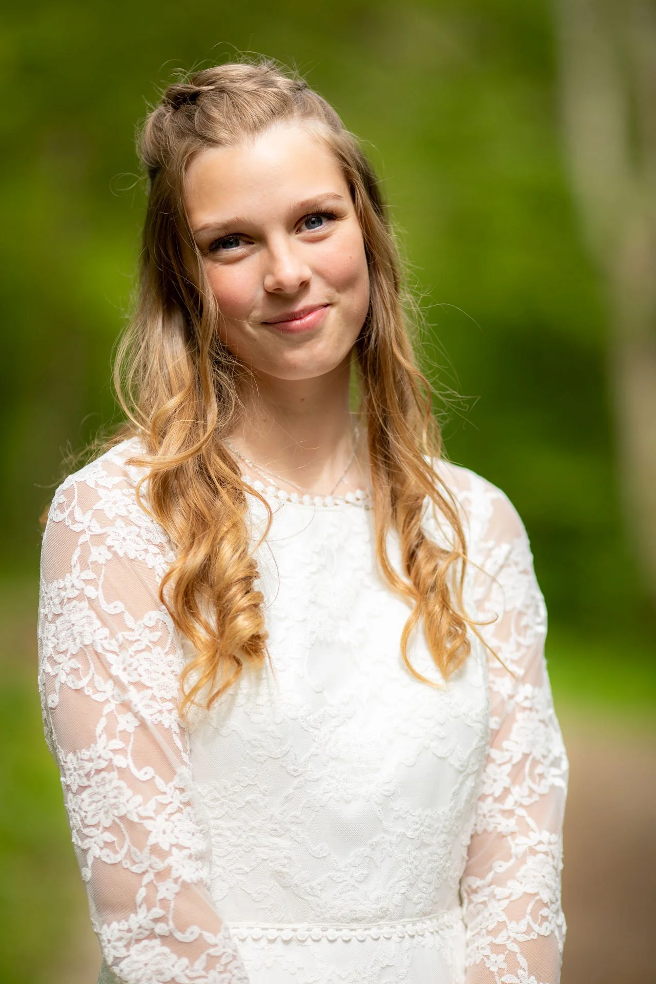 Portræt af en ung kvinde i hvid kjole med blondeærmer, udendørs med grøn baggrund.